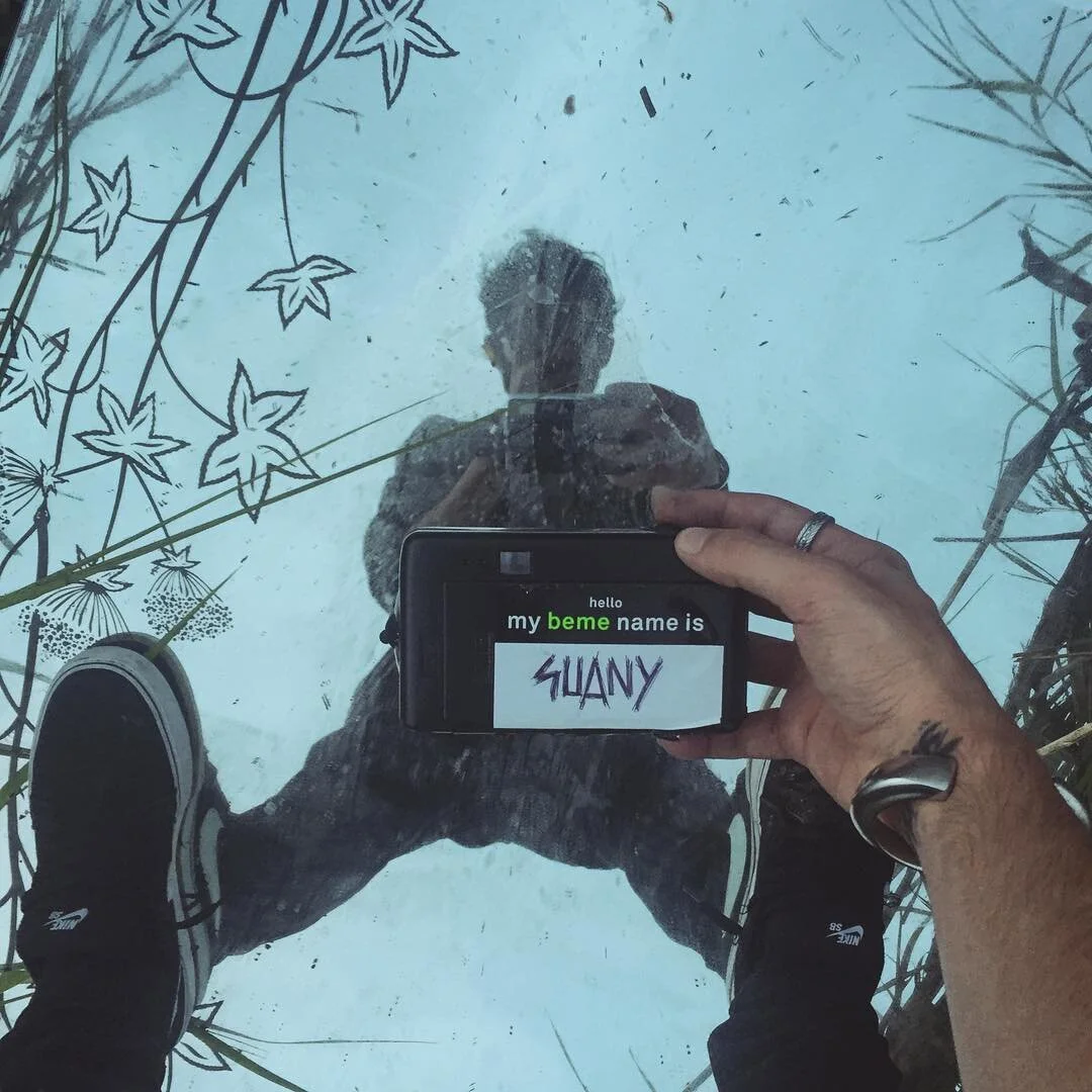Person taking a photo of their reflection on a glass surface with floral and leaf patterns. The person's legs and shoes are visible, as well as their hand holding a phone displaying the name 'SUANY' on a screen that says 'hello my beme name is'.