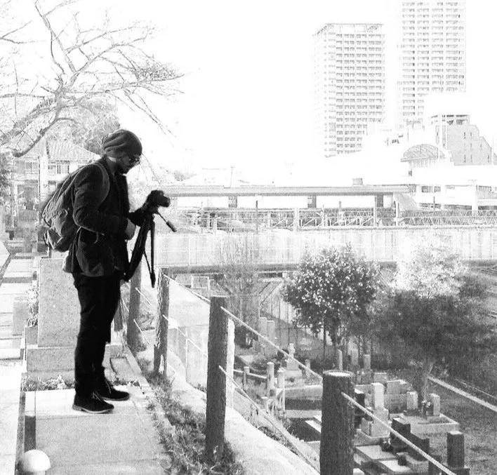 A person standing on a ledge with a camera, overlooking a cityscape with trees, buildings, and an elevated walkway or bridge.