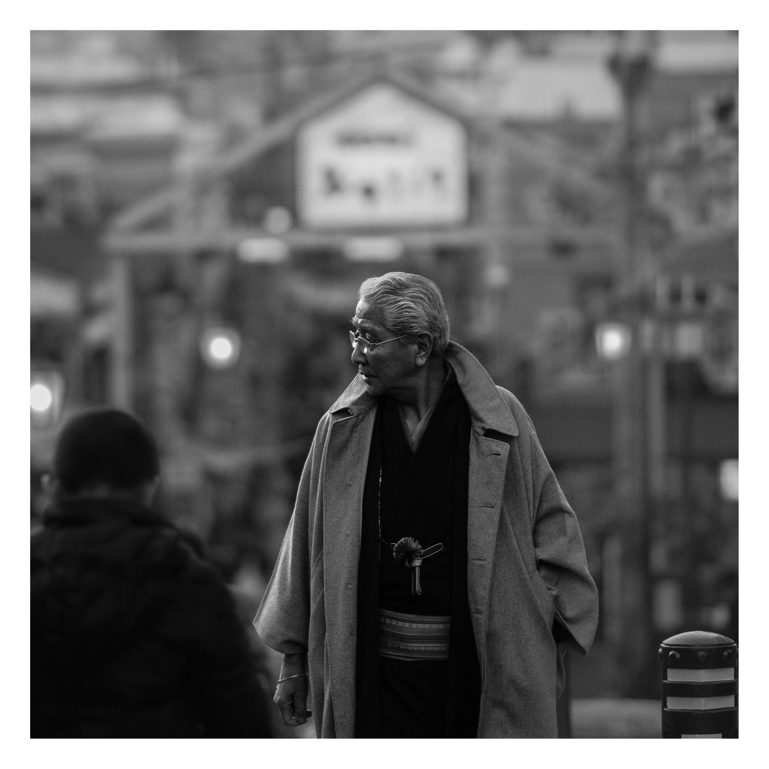 A black-and-white photograph of an older man with glasses, dressed in a coat, walking outdoors in a city scene with blurred people and buildings in the background.