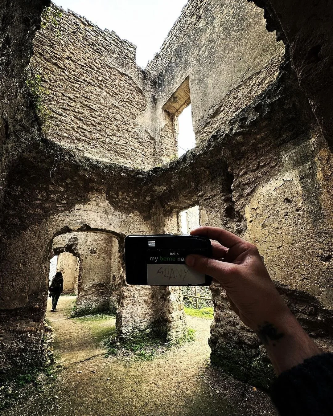A person taking a photo inside an old, partially ruined stone building with arched doorways and windows. The photographer's hand and phone are visible in the foreground, and another person is visible walking through a doorway in the background.