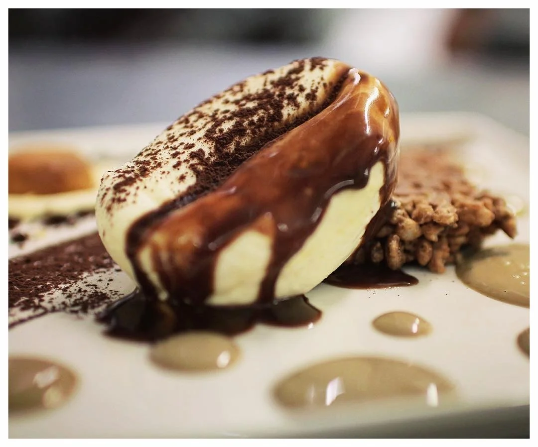 Close-up of a banana split dessert with vanilla ice cream, chocolate syrup, cocoa powder, and crushed nuts on a white plate.