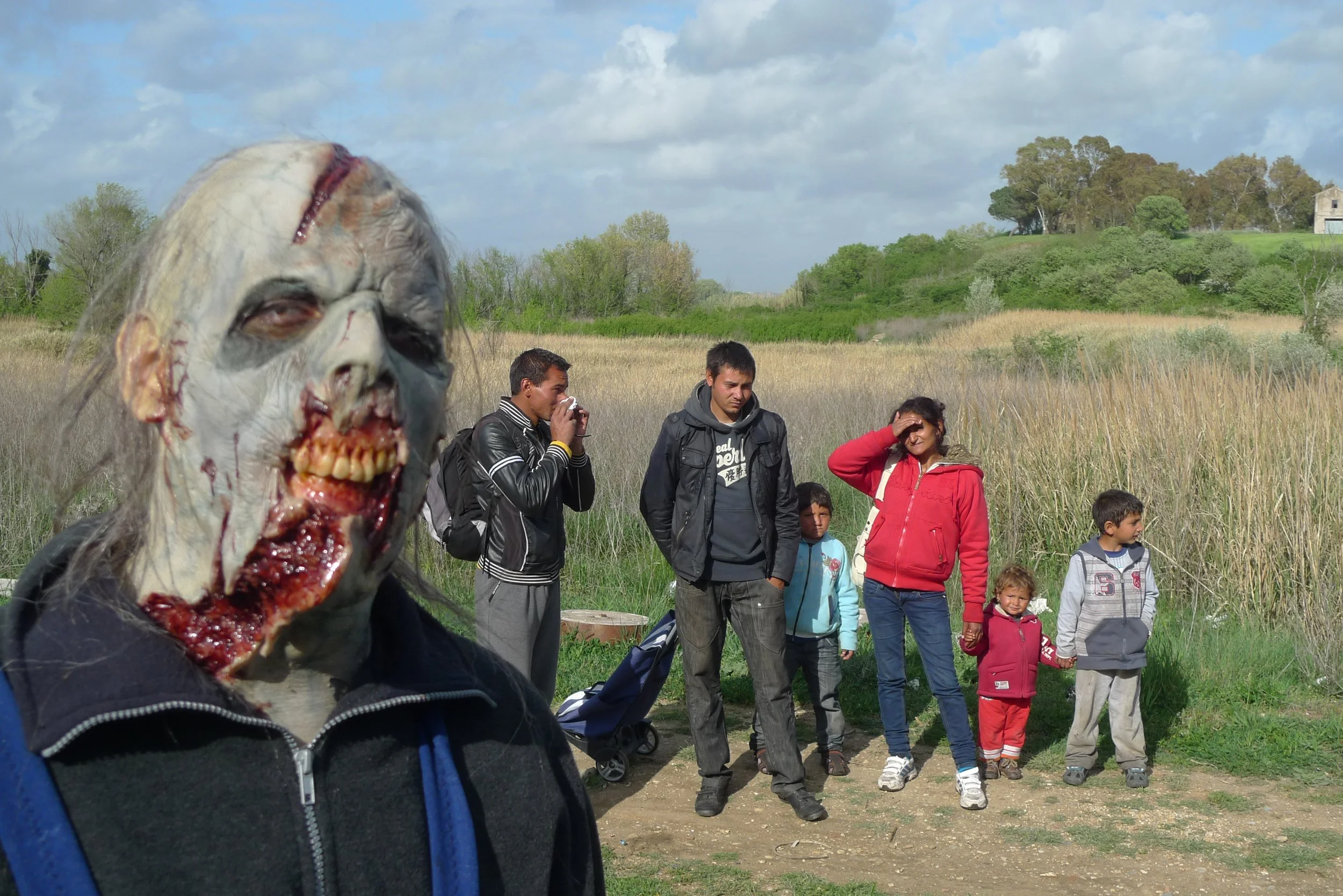 A group of people, including children, standing outdoors in a grassy field with trees and a cloudy sky in the background, with a person wearing a zombie mask in the foreground.