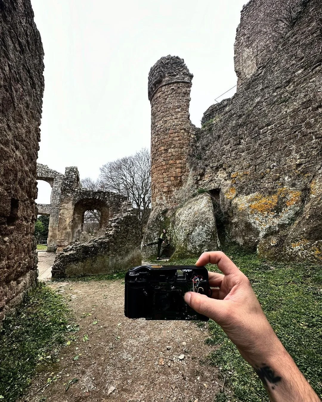 A person taking a photograph of ancient stone ruins with a digital camera, showing a tall, rounded tower and stone archways surrounded by moss and overgrown vegetation.