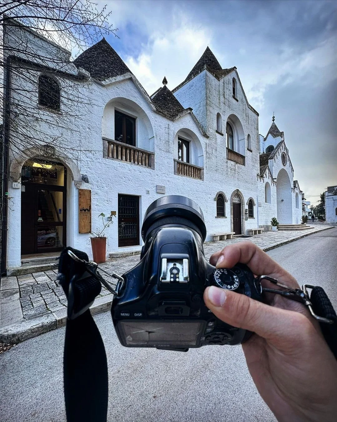 Person holding a camera in front of a white historic building with arched windows and pointed roofs, on a quiet street, under a partly cloudy sky.
