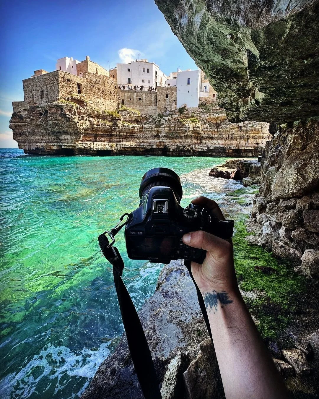 A person holding a camera on a rocky shoreline, capturing a view of white buildings on a cliff overlooking turquoise water and a blue sky.