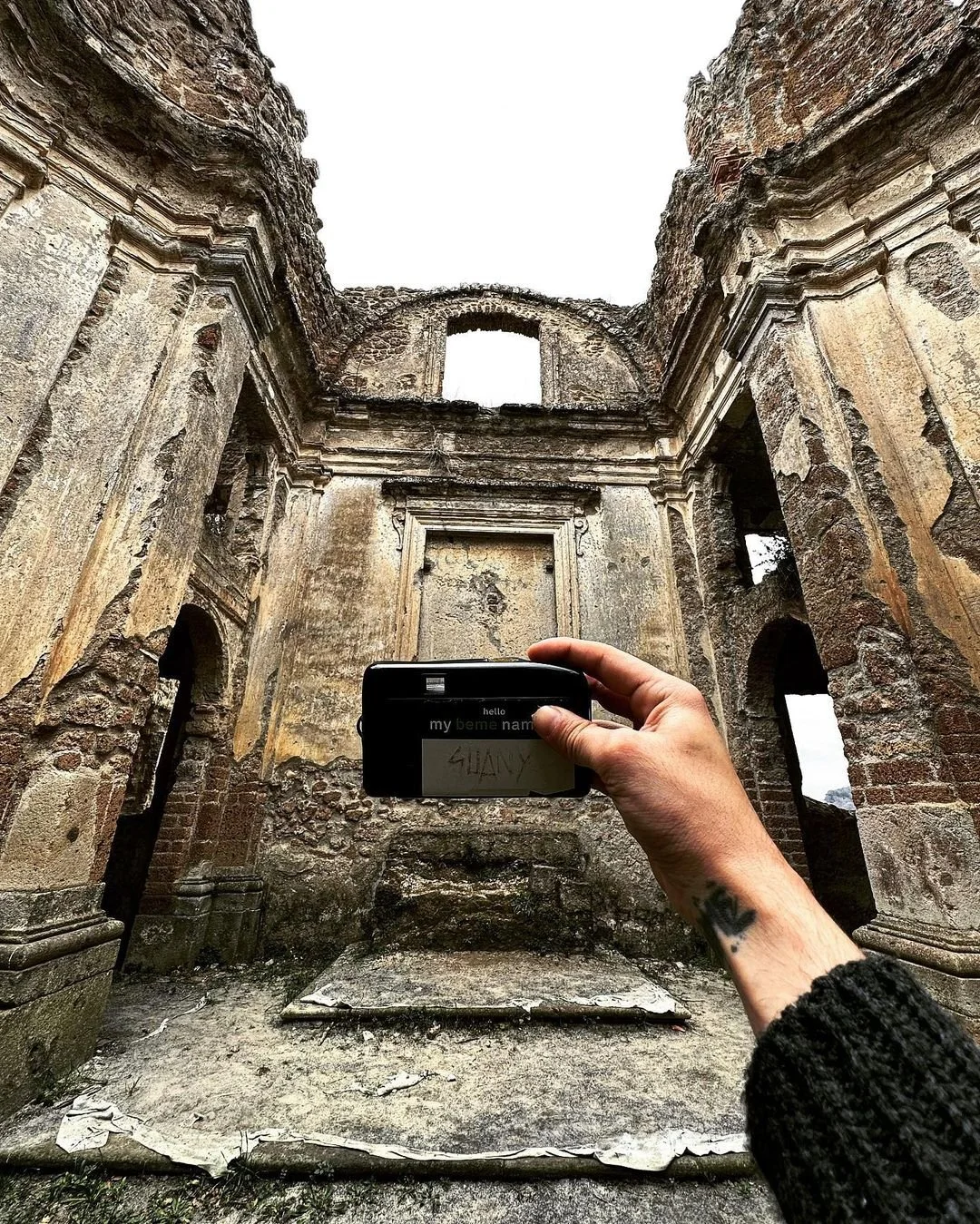 Person holding a camera in front of the ruins of an old, weathered stone building with tall walls and archways.
