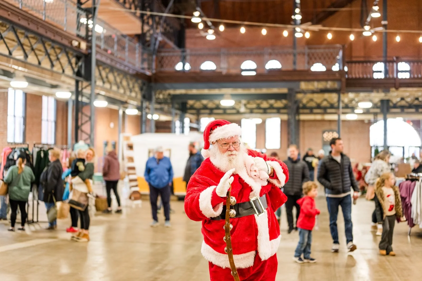 Santa greeting families at the Illinois Local Holiday Market on Small BSanta Claus in a red suit with black boots and a white beard taking a photo with a person in a Sonic the Hedgehog costume inside a large indoor venue with industrial architecture.