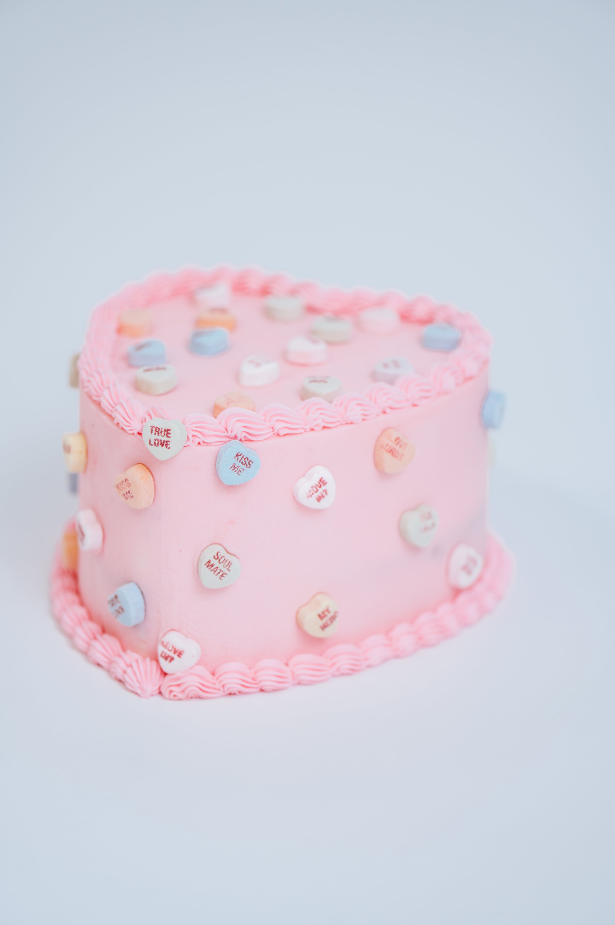 A pink heart shaped cake with conversation hearts around the outside 