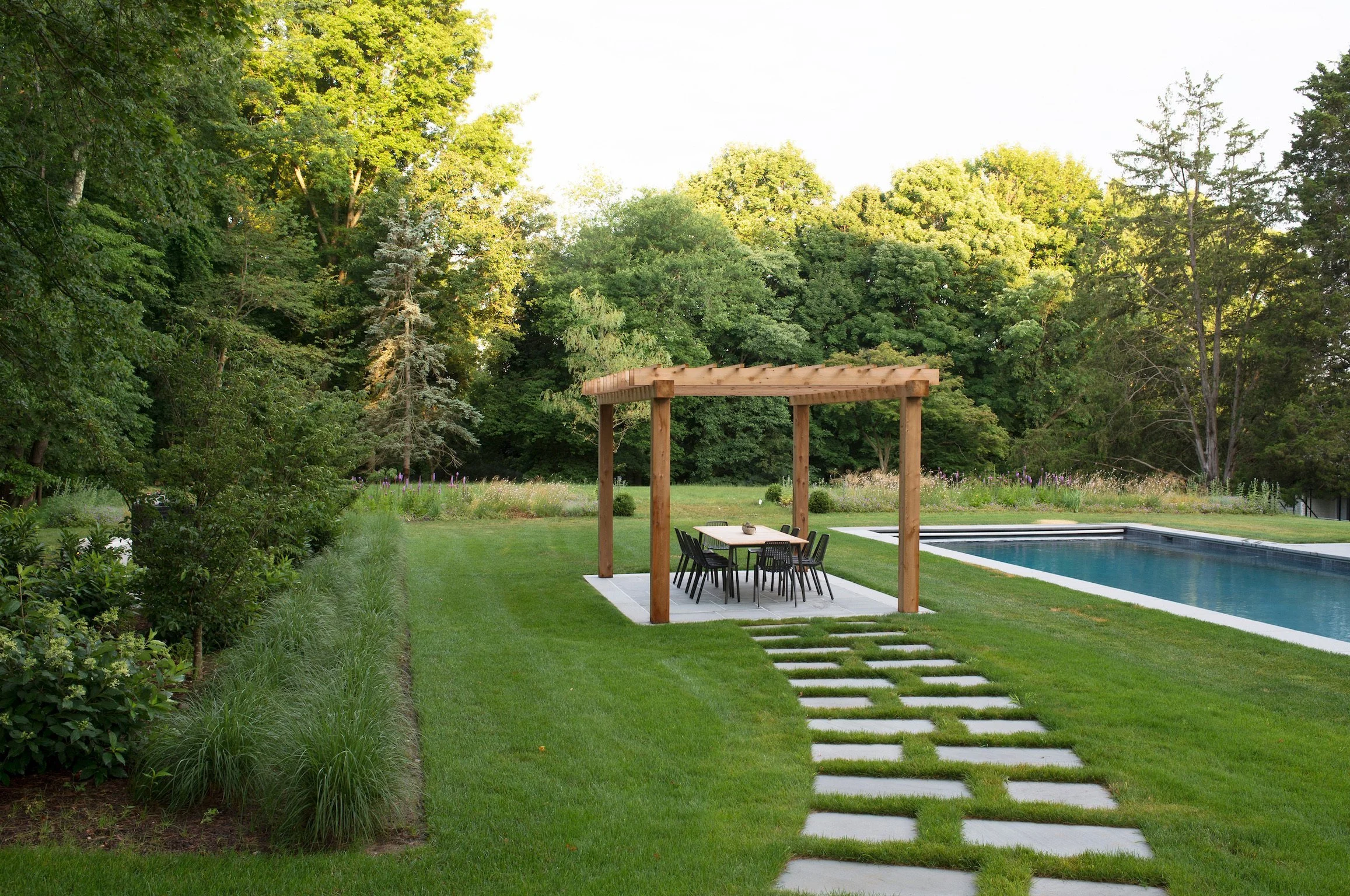 A backyard scene featuring a swimming pool, grassy lawn, stone stepping path, a wooden pergola over an outdoor dining table and chairs, with tall green trees in the background.