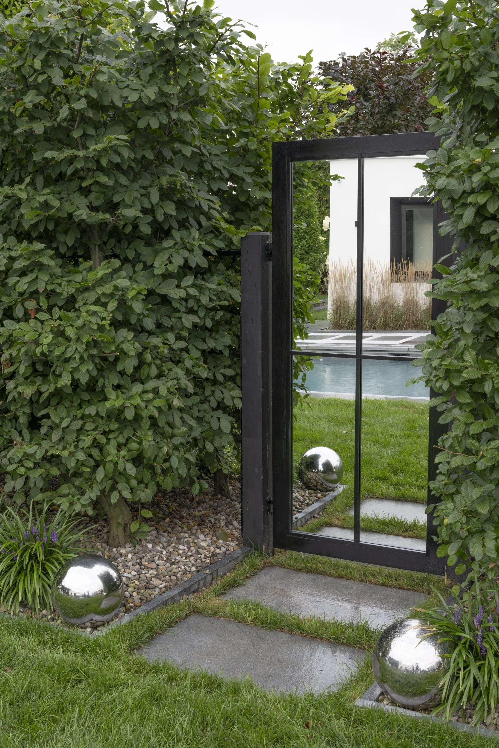 A modern house entrance with a glass door, surrounded by lush green bushes, ornamental grass, and decorative silver spheres.
