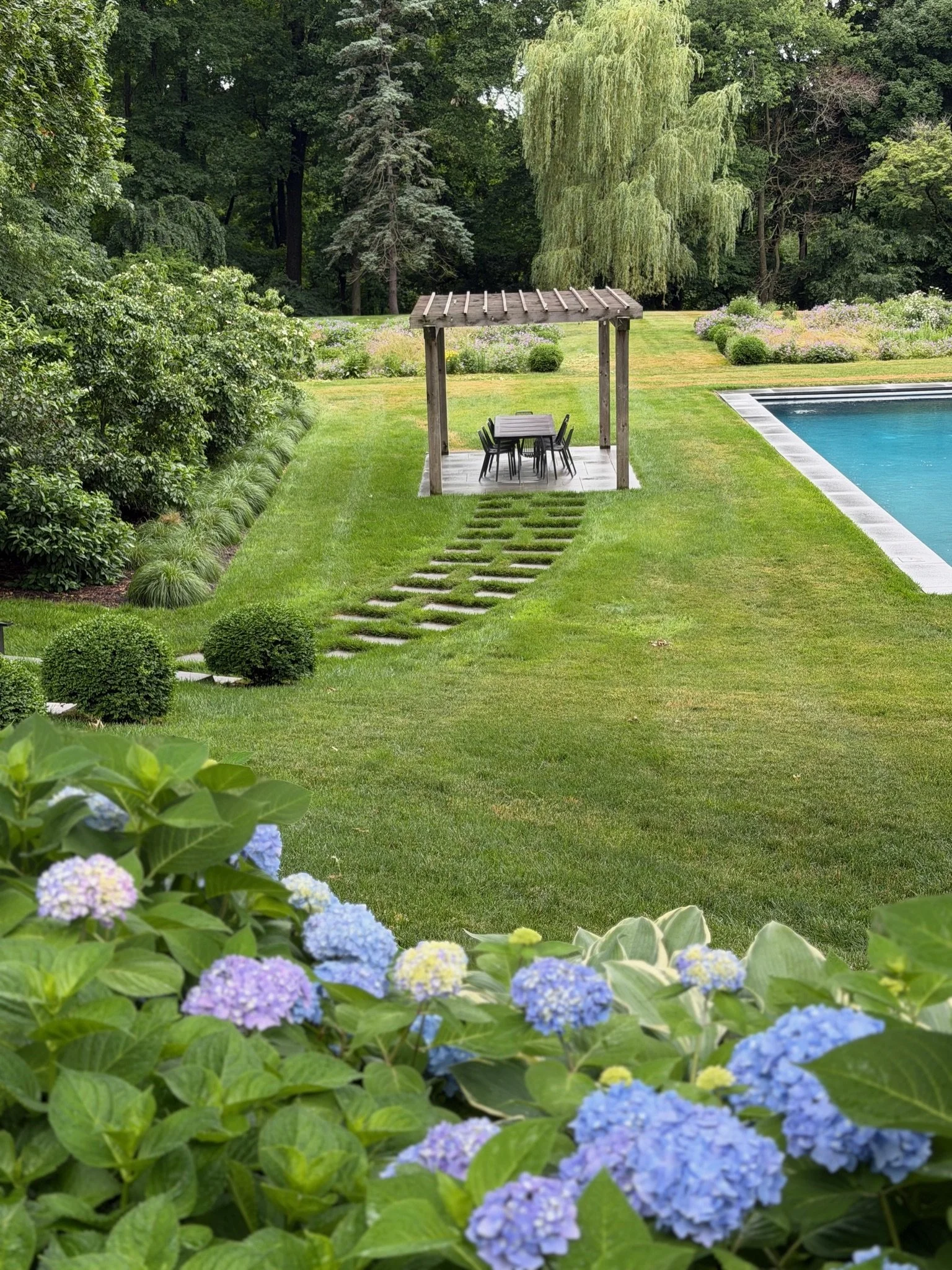 A backyard garden with a lawn, a wooden pergola with a table and chairs underneath, a swimming pool, and a variety of trees and bushes in the background.