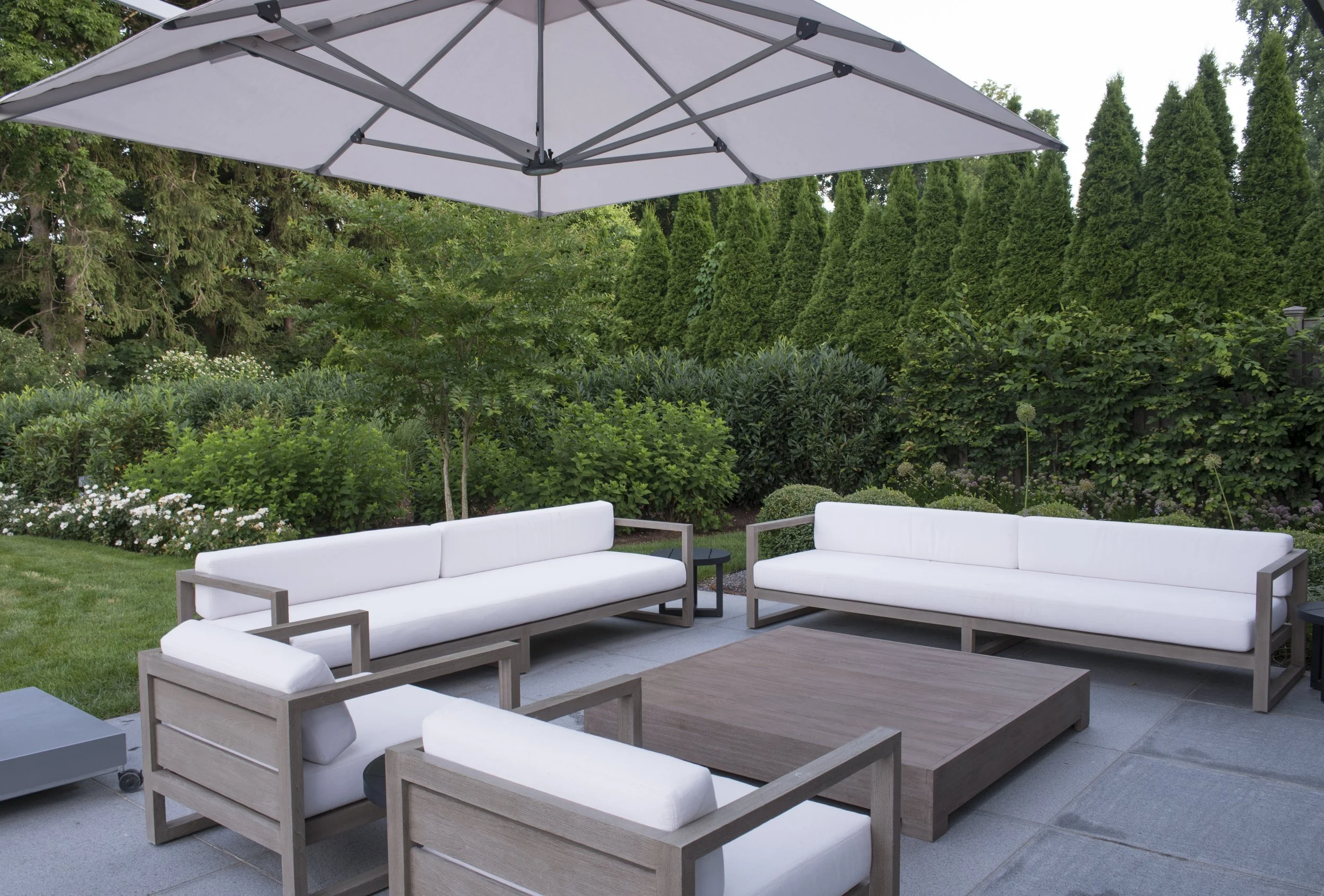 White cushioned lounge seating under a large umbrella, screened by evergreens