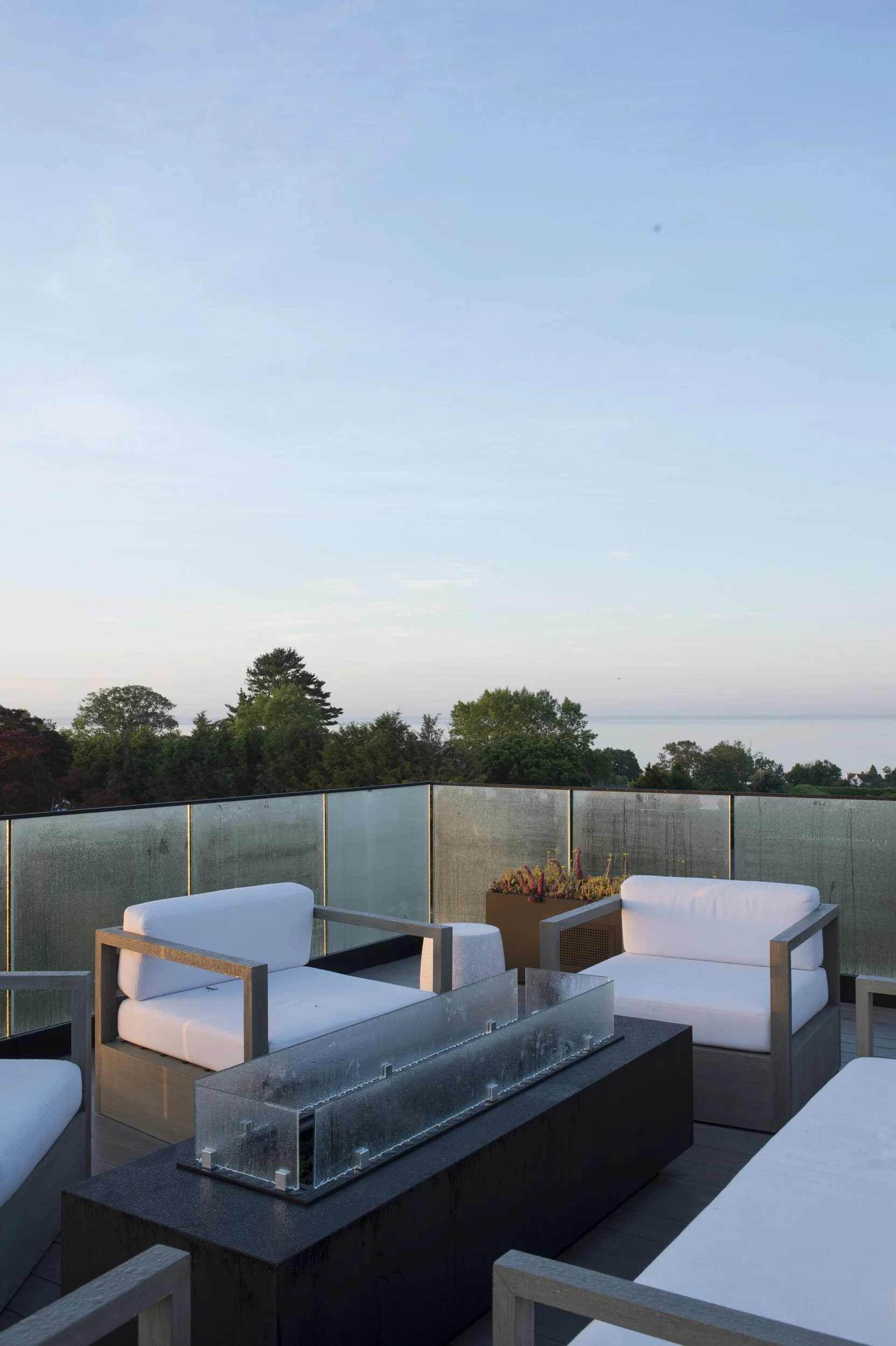 Roof deck with white lounge seating around a glass fire table, overlooking the water