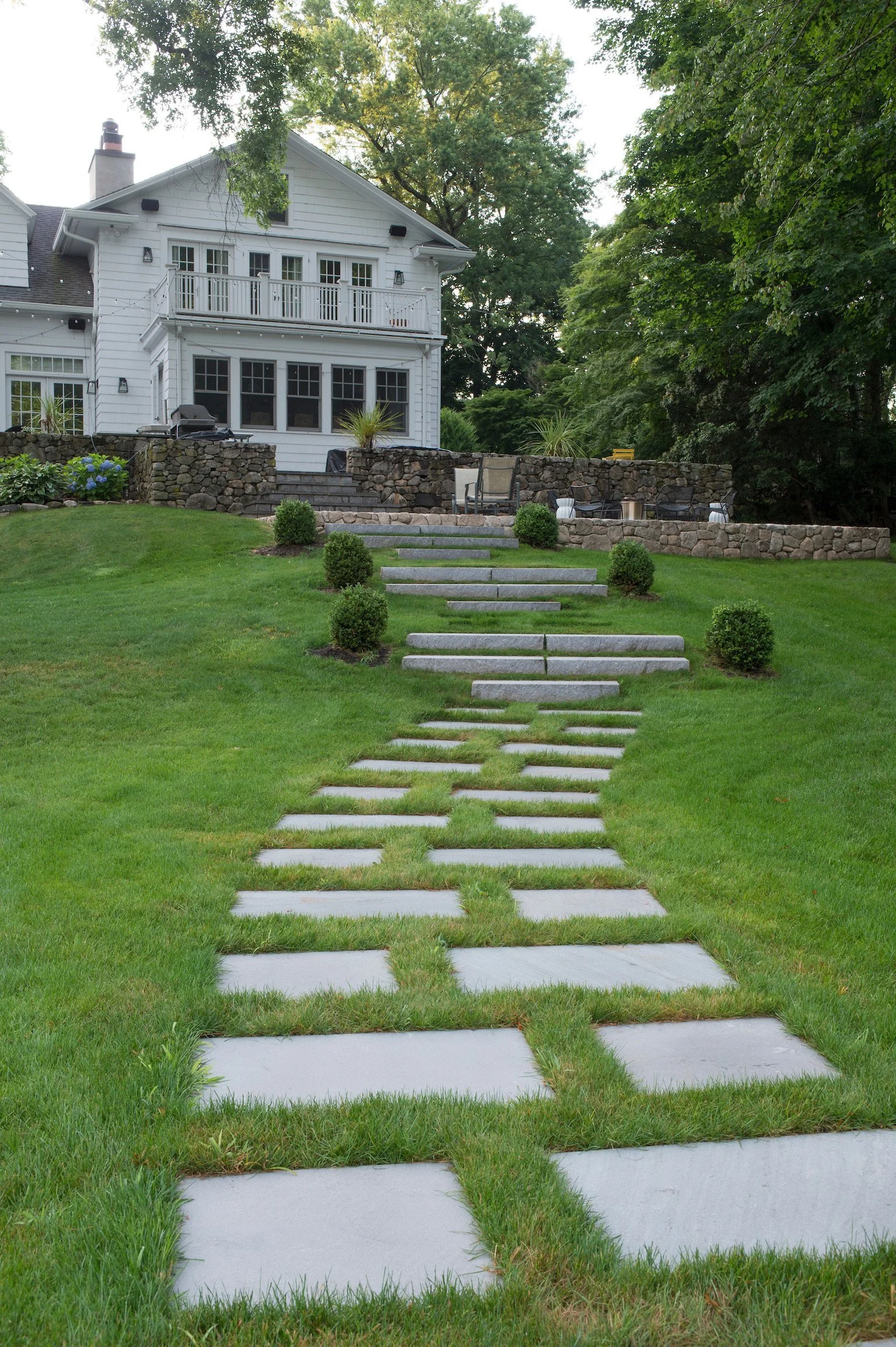 A lawn with a stone pathway leading up to a stone patio with outdoor furniture in front of a large white house with multiple windows and a balcony, surrounded by trees.