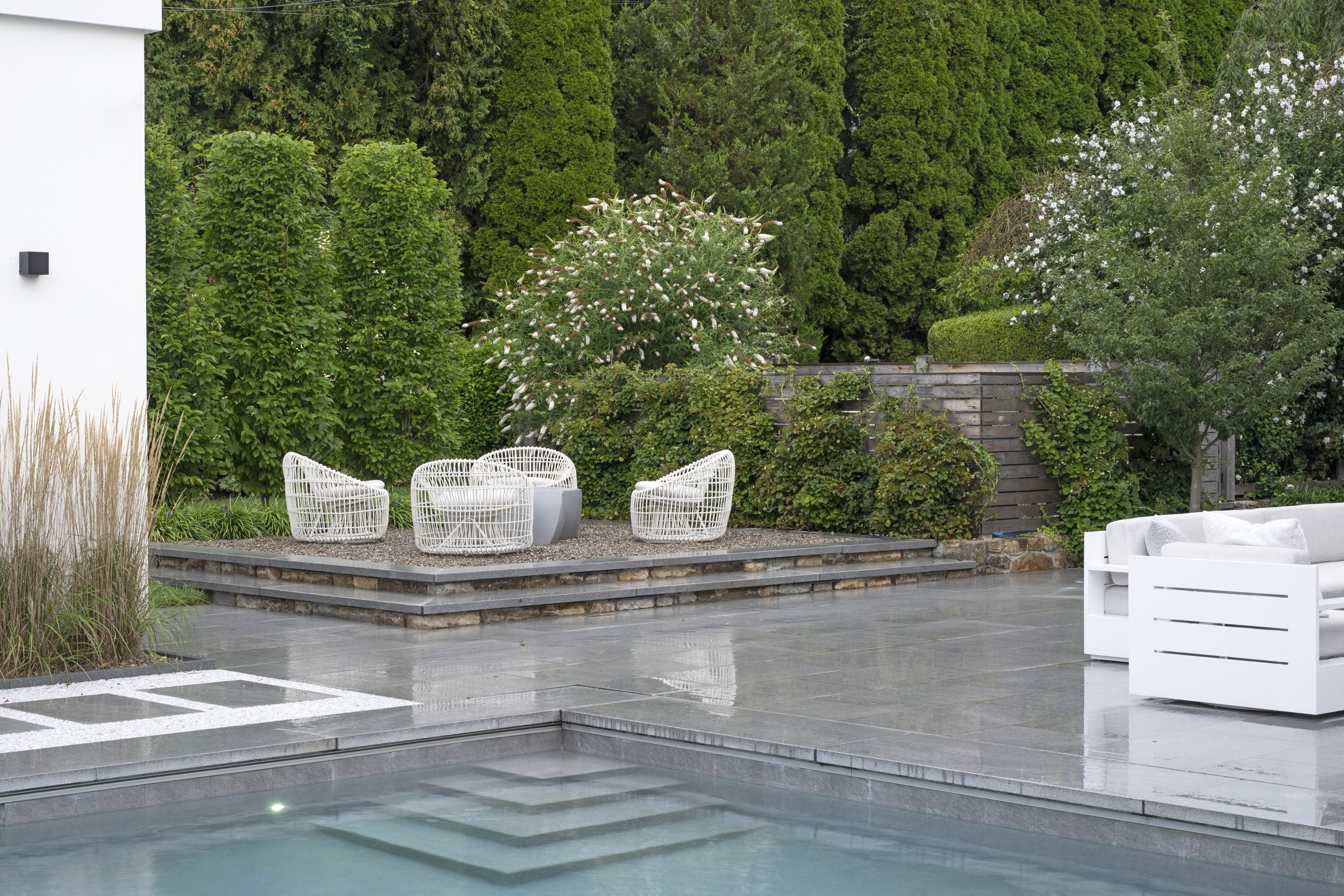 A modern outdoor patio with white woven chairs, a small round table, and lush green trees and bushes in the background.