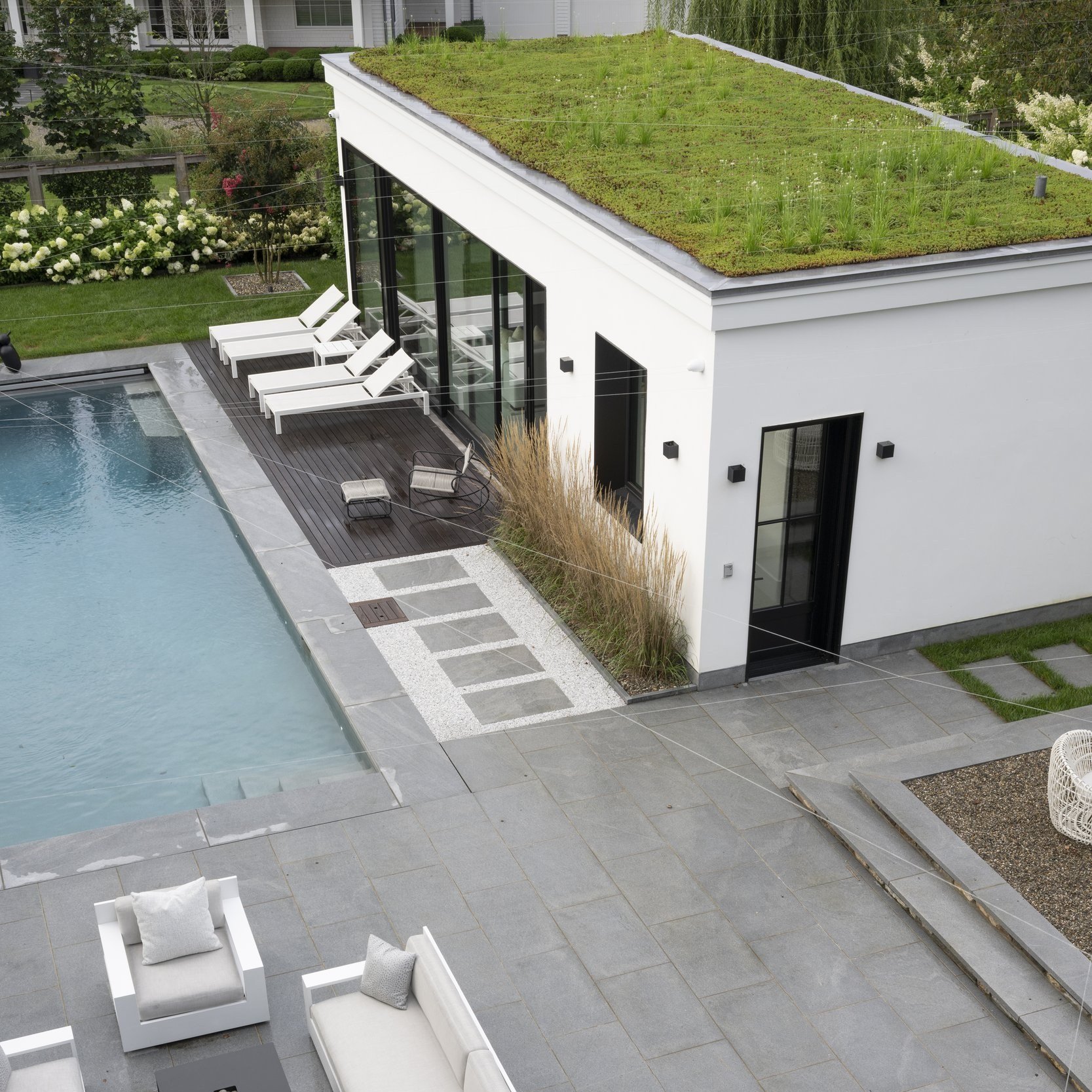 View from above of a pool house with a planted green roof, a long swimming pool and a stone terrace with white outdoor seating.
