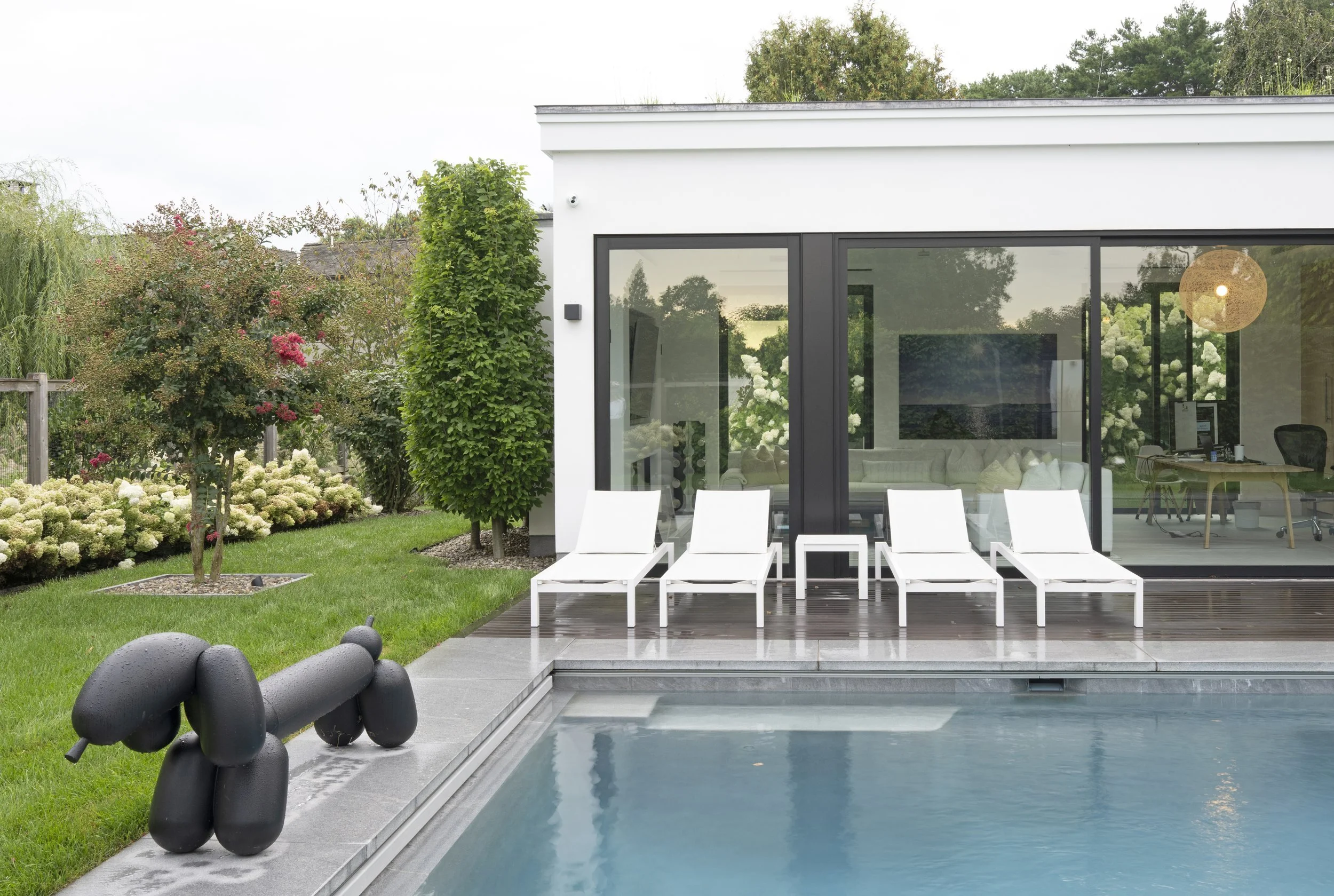 Modern house with large glass windows, outdoor pool, white lounge chairs, landscaped garden with flowering bushes and trees, black abstract sculpture near the pool, and patio area.