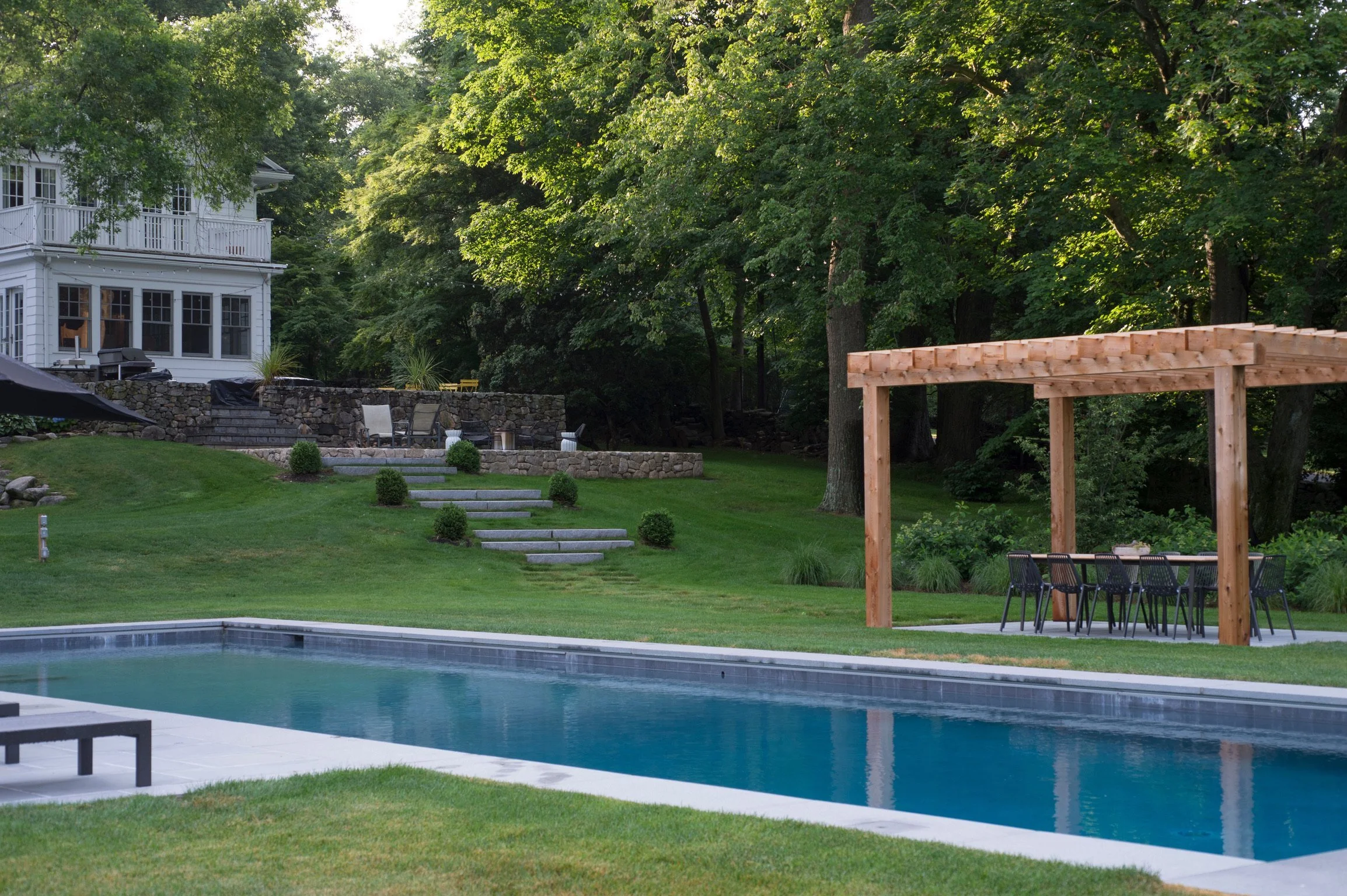 Residential backyard with a swimming pool, outdoor furniture, a wooden pergola, a stone patio, landscaped grass, and a white house surrounded by mature trees in the background.