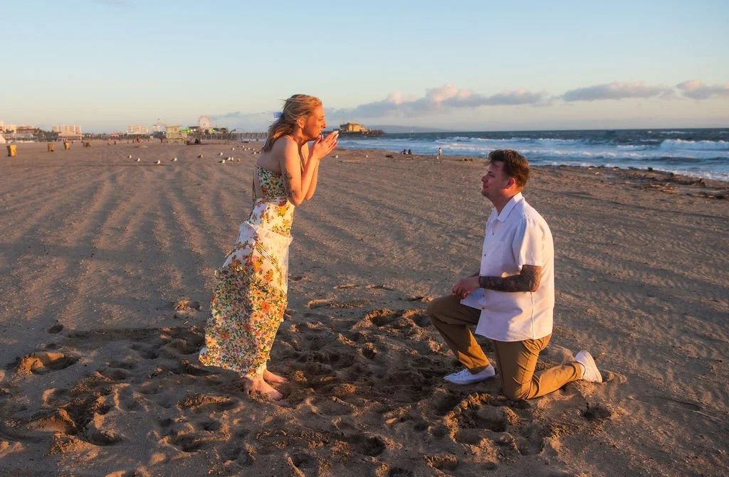 A man kneeling on the beach, proposing to a woman who looks surprised, near the ocean with a city skyline in the background.