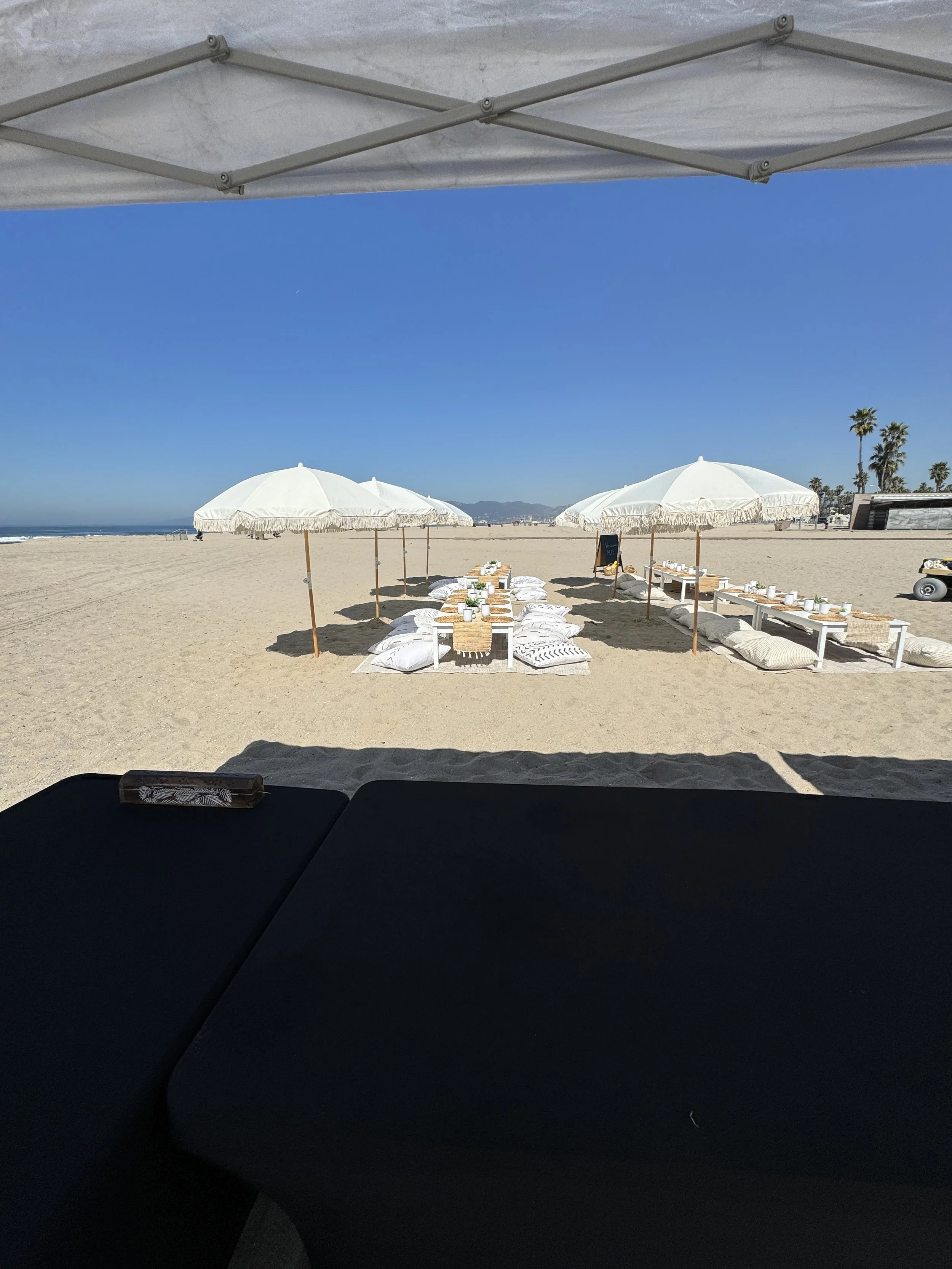 Empty beach with white umbrellas and low tables set for dining, under a canopy on sunny day.