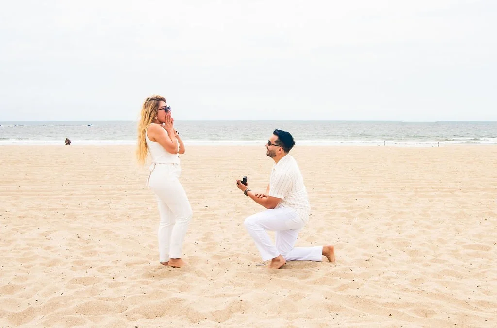 A man is proposing to a woman on the beach, with the woman showing surprise and happiness.