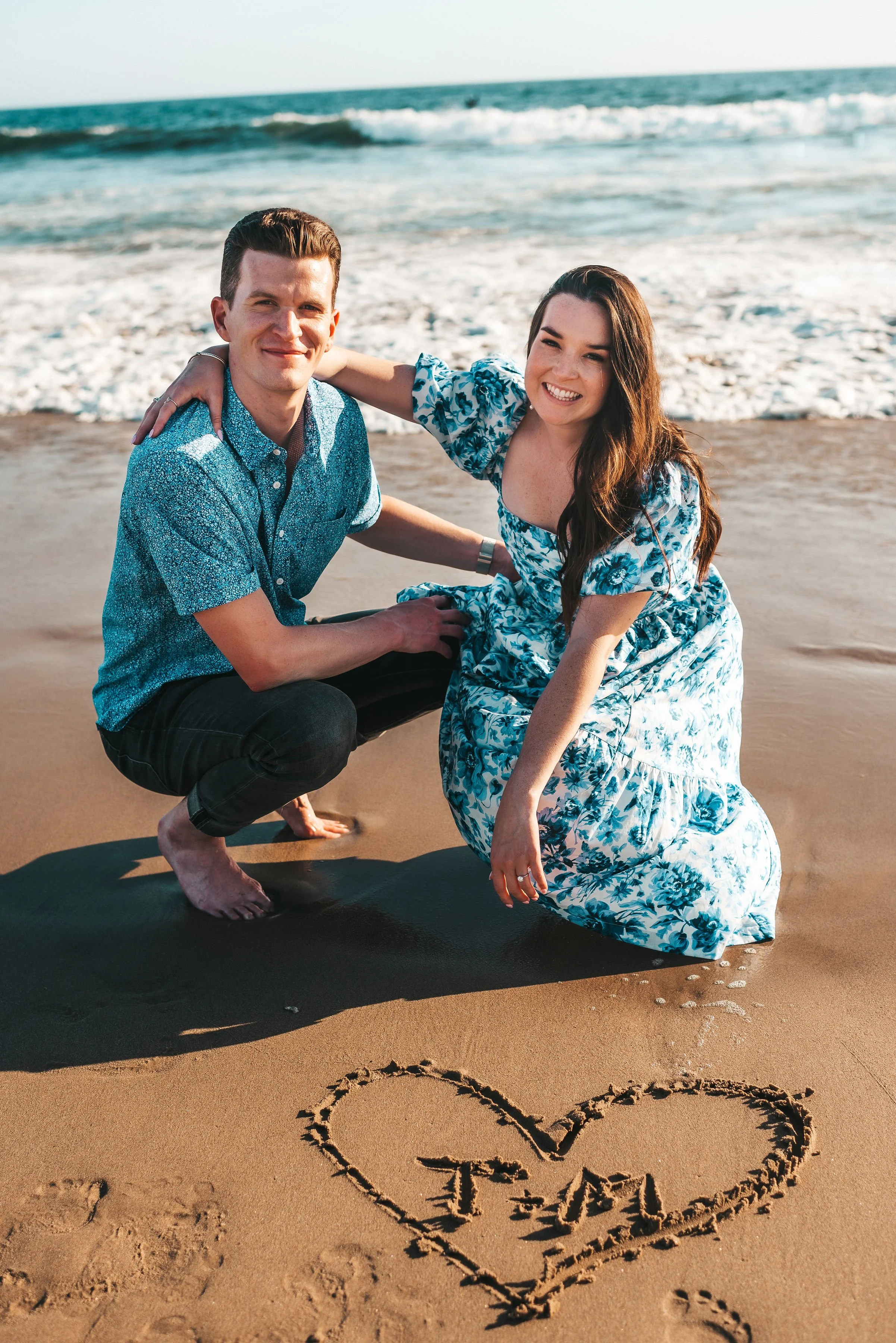 A couple on the beach, with the man squatting and the woman kneeling, smiling, next to a heart drawn in the sand with their initials T and A inside.