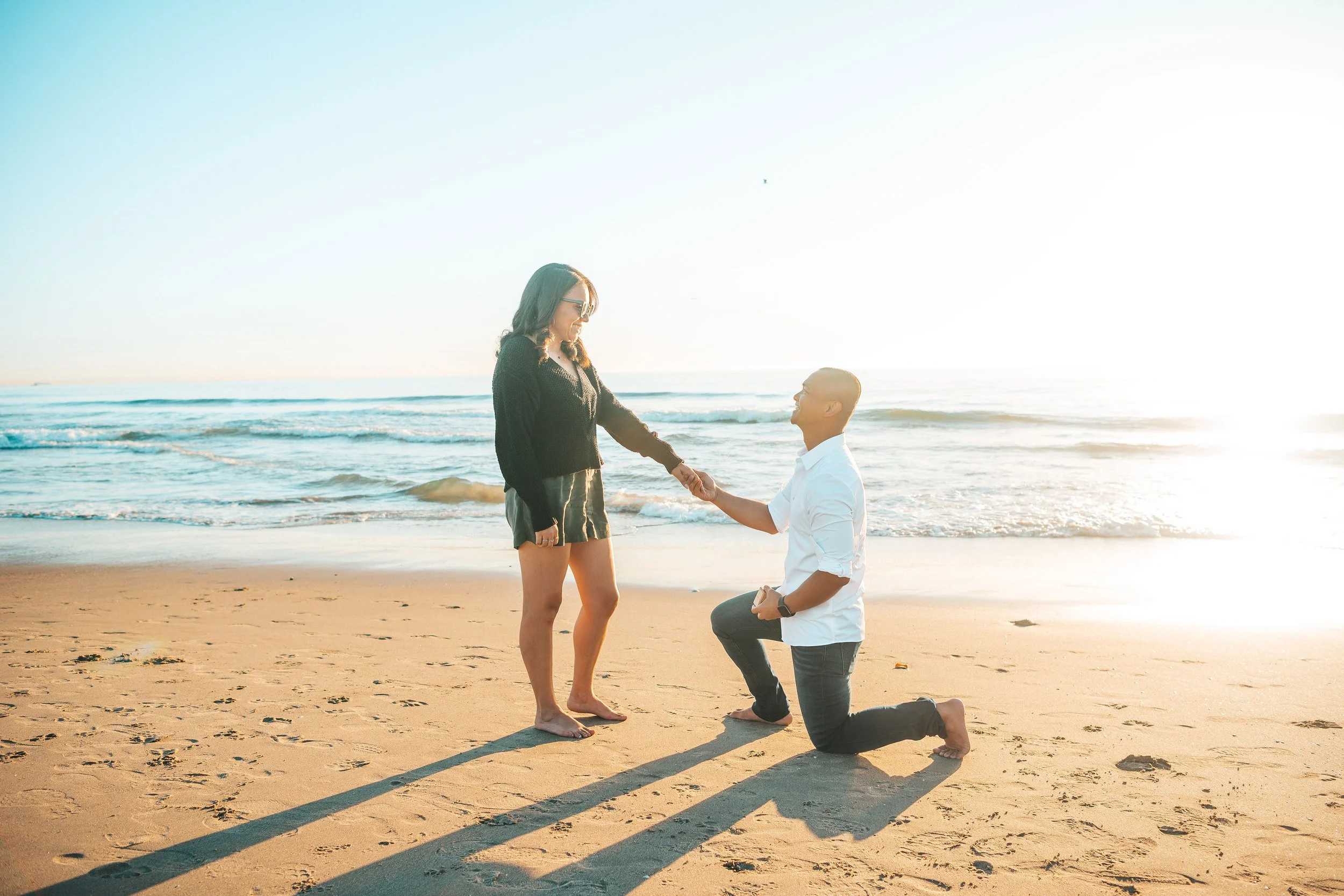 A man proposes to a woman on the beach at sunset, with the man kneeling on one knee and holding the woman's hand.
