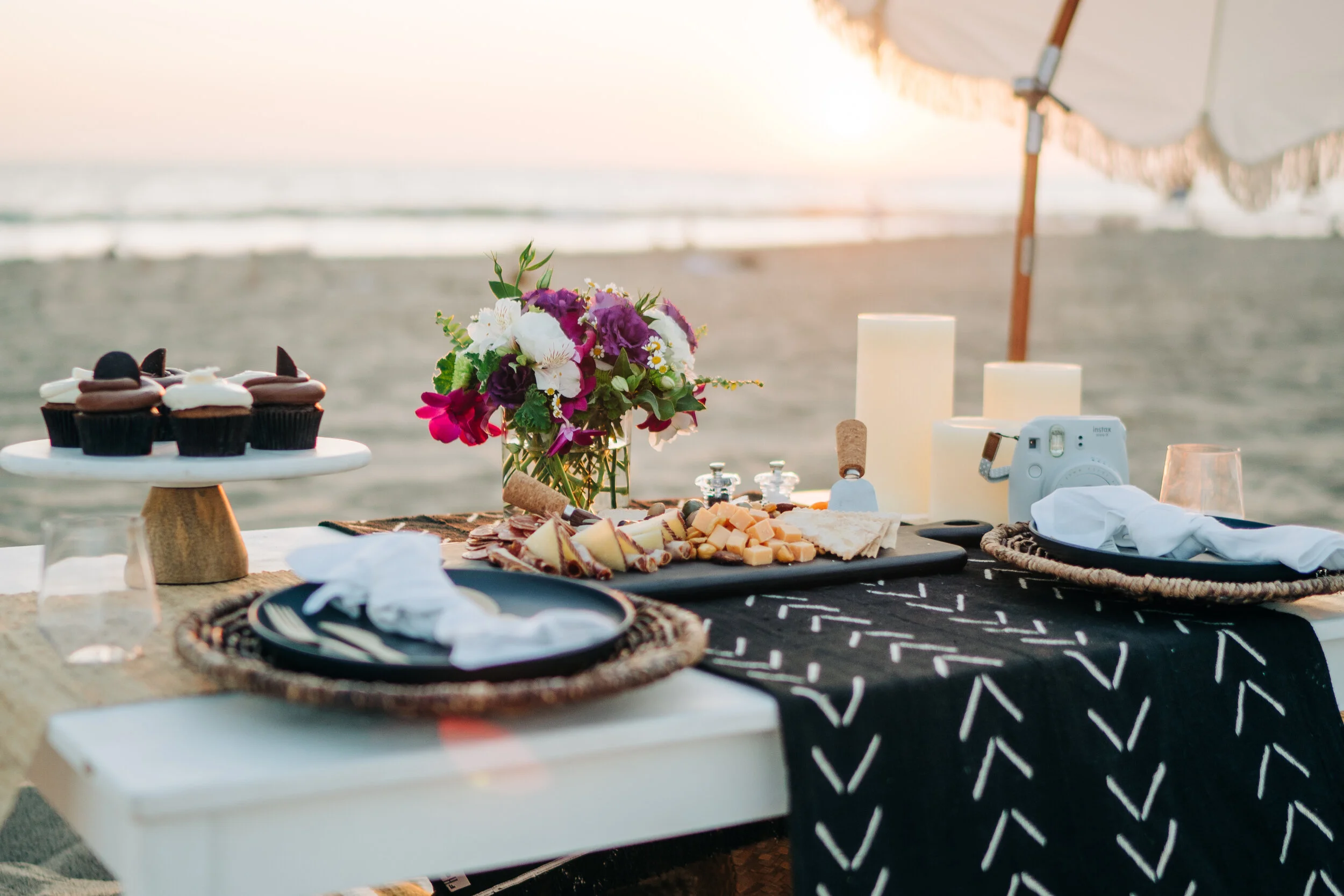 Beachside table set with cupcakes, cheese, and flowers, under an umbrella at sunset.