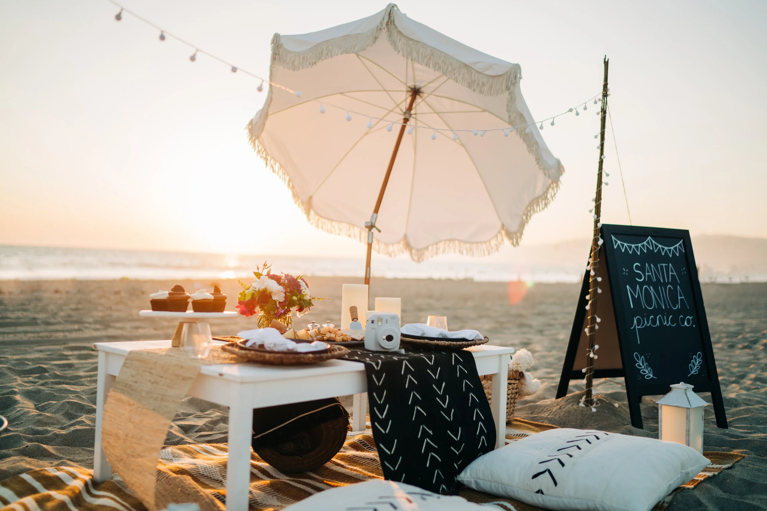 Beach picnic setup on sand with a white umbrella, black chalkboard sign reading 'Santa Monica picnic co.', a low table with food, flowers, and a camera, and pillows on the sand during sunset.