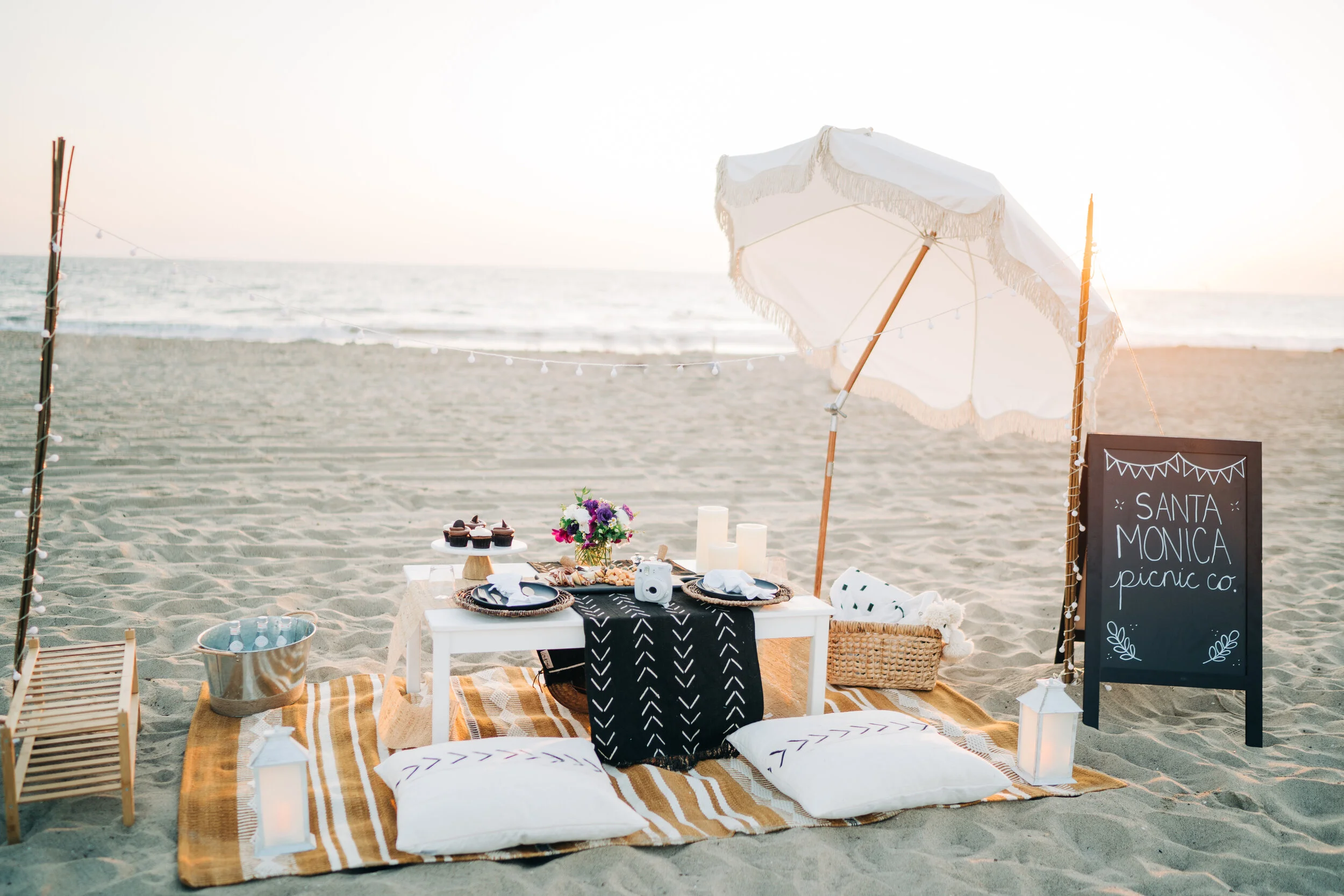 Beach picnic setup with a small table, pillows, flowers, cupcakes, and a chalkboard sign that reads 'Santa Monica picnic co' on sand, with ocean in the background and a white beach umbrella.