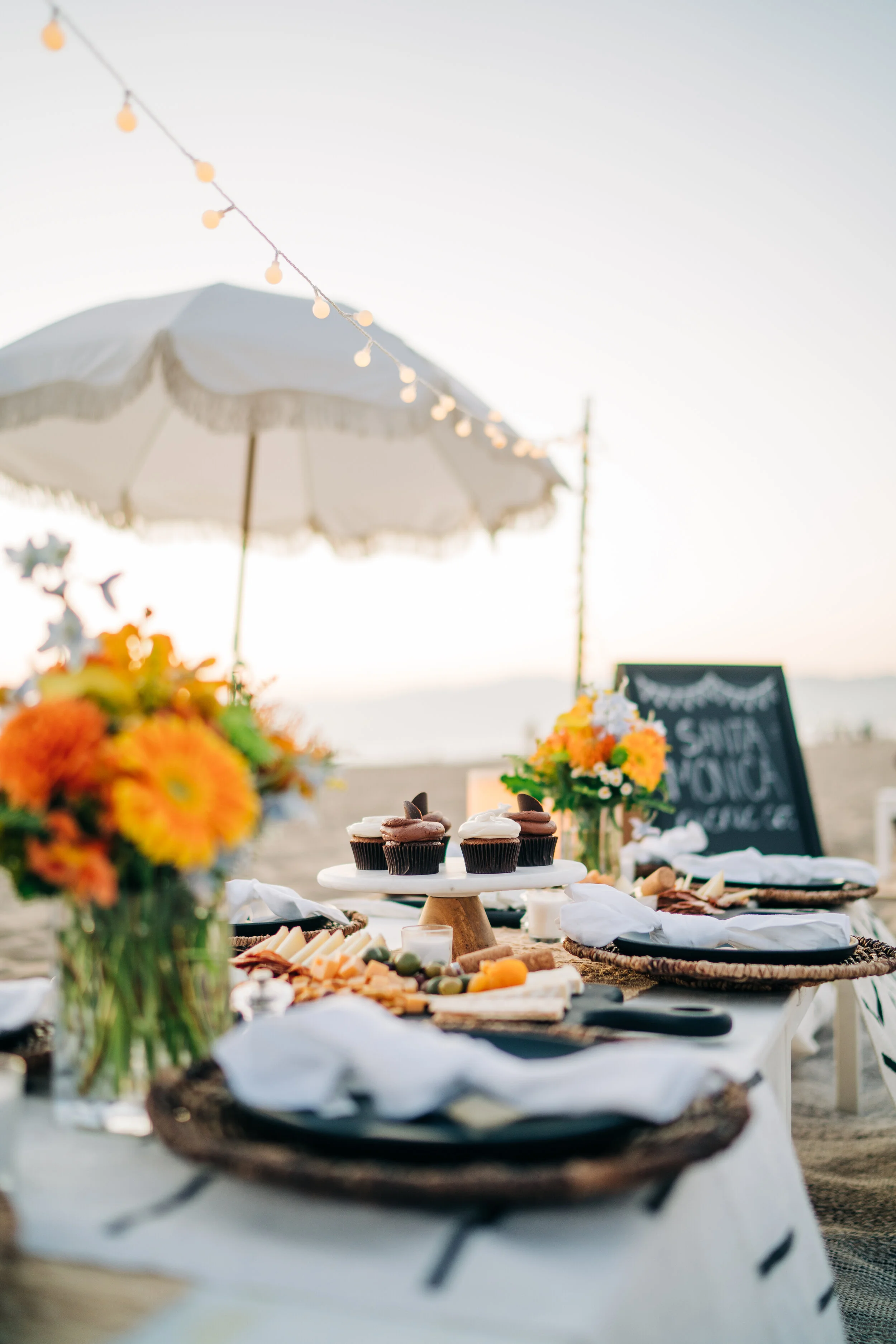 A beachside picnic setup with a table of food and flowers, a sun umbrella, and string lights at sunset.