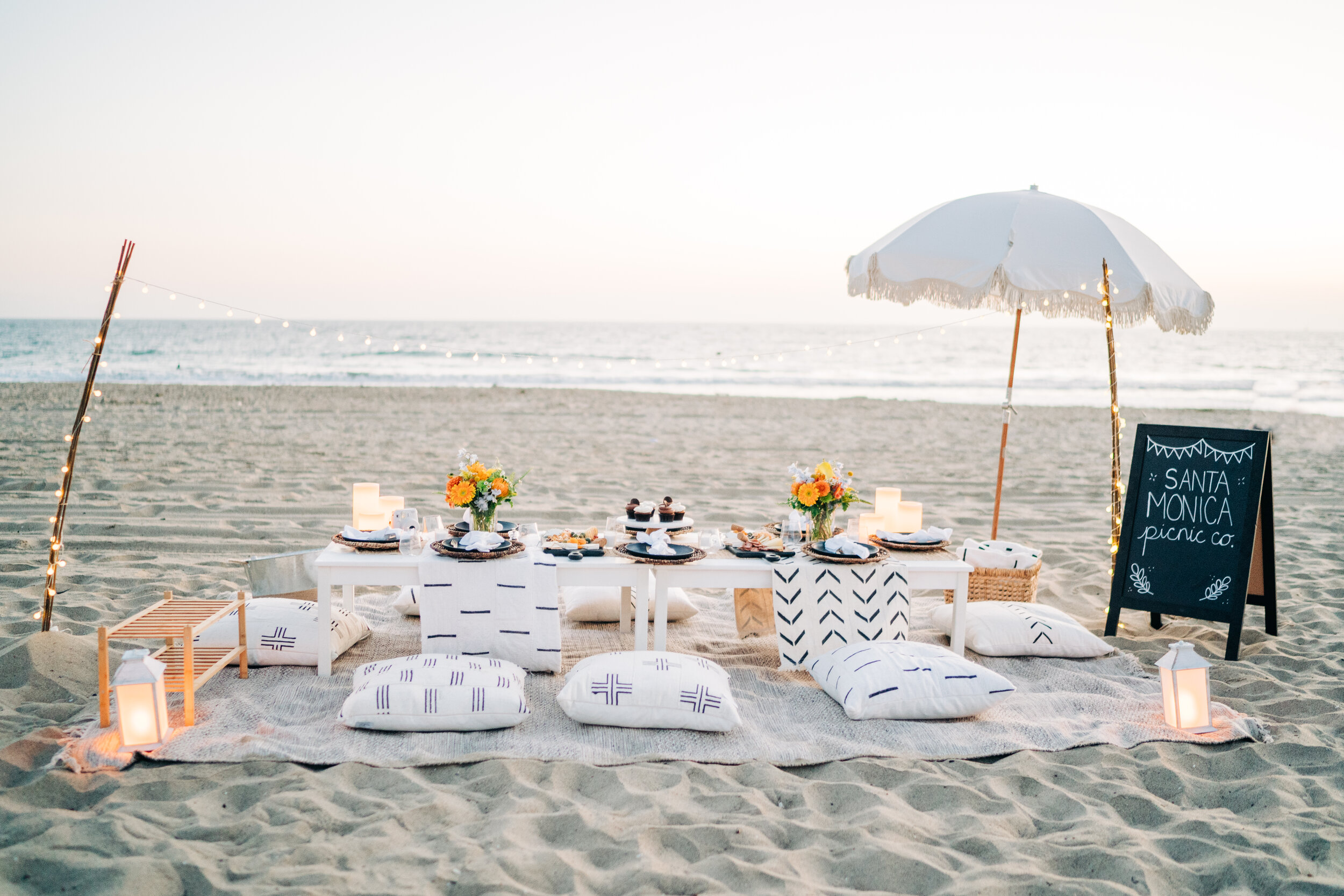 Beachside picnic setup with a low white table, cushions on sand, flowers, candles, cupcakes, and a chalkboard sign reading 'Santa Monica Picnic Co.' under a white umbrella, with the ocean and sky in the background.