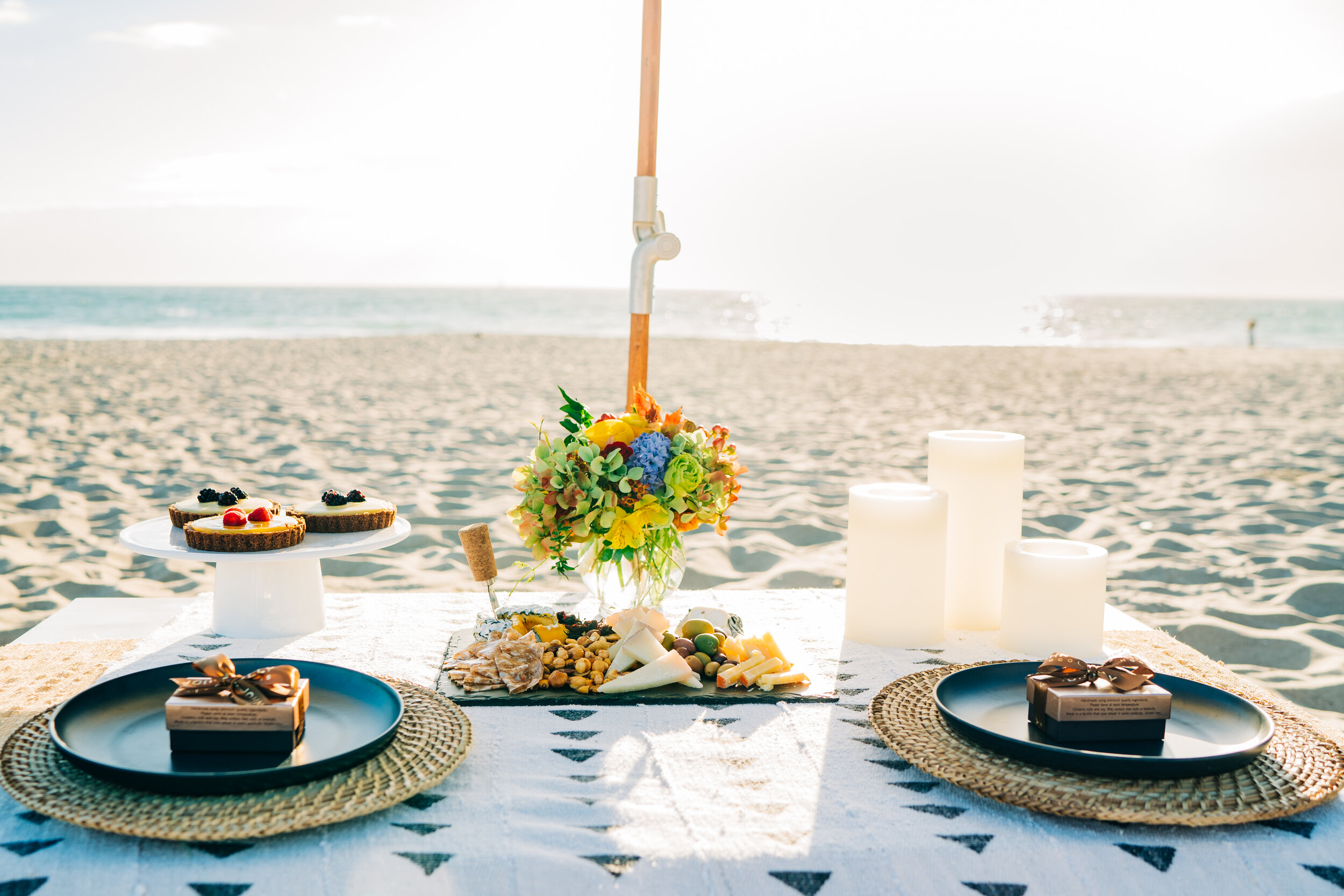 Beachside picnic table with cheese, crackers, fruit, flowers, and desserts on a white tablecloth, set against an ocean background with candles.