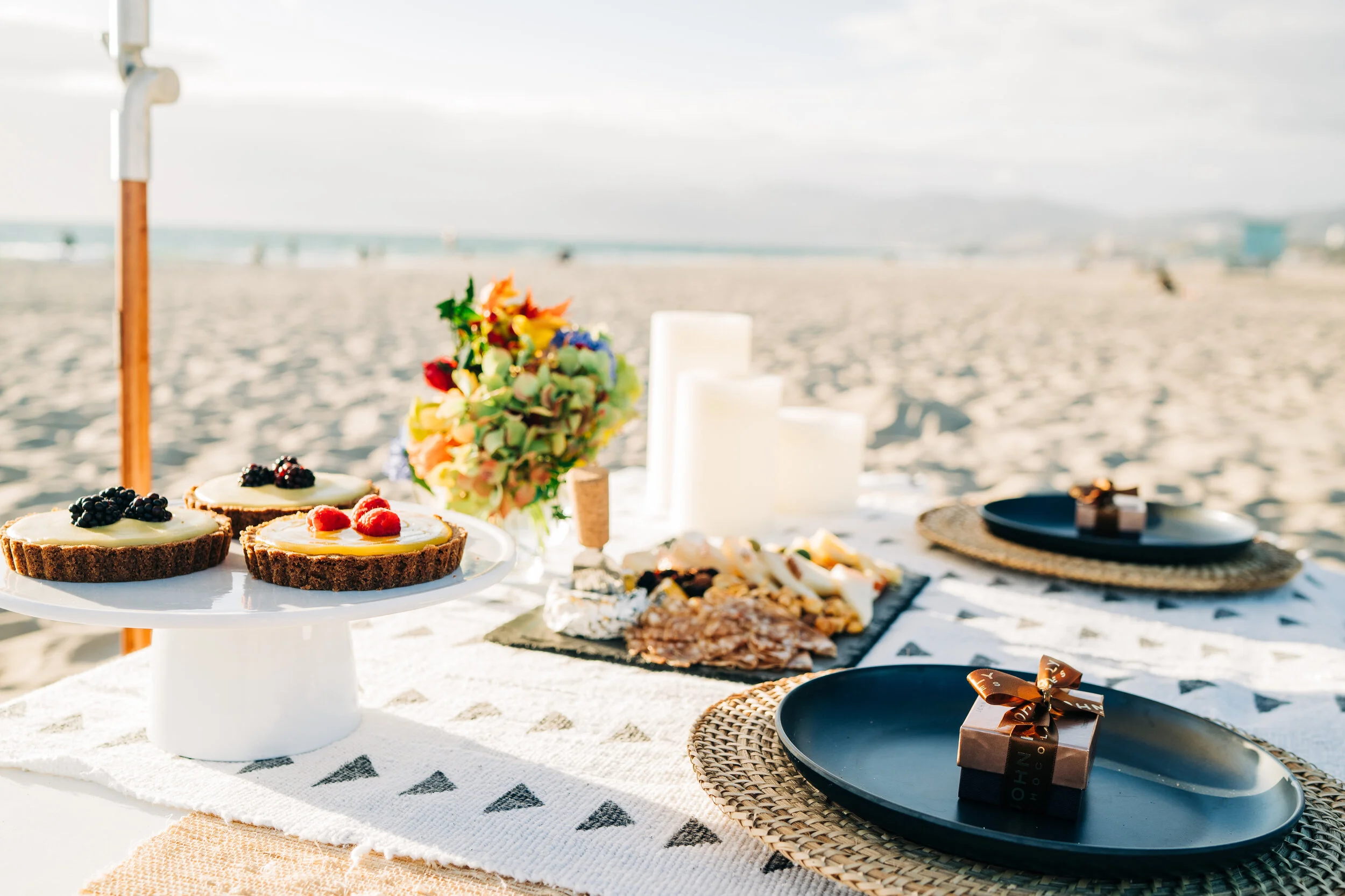 Beachside picnic setup with fruit-topped desserts, cheese platter, flowers, candles, and wrapped gifts on a table with a sandy beach and ocean in the background.
