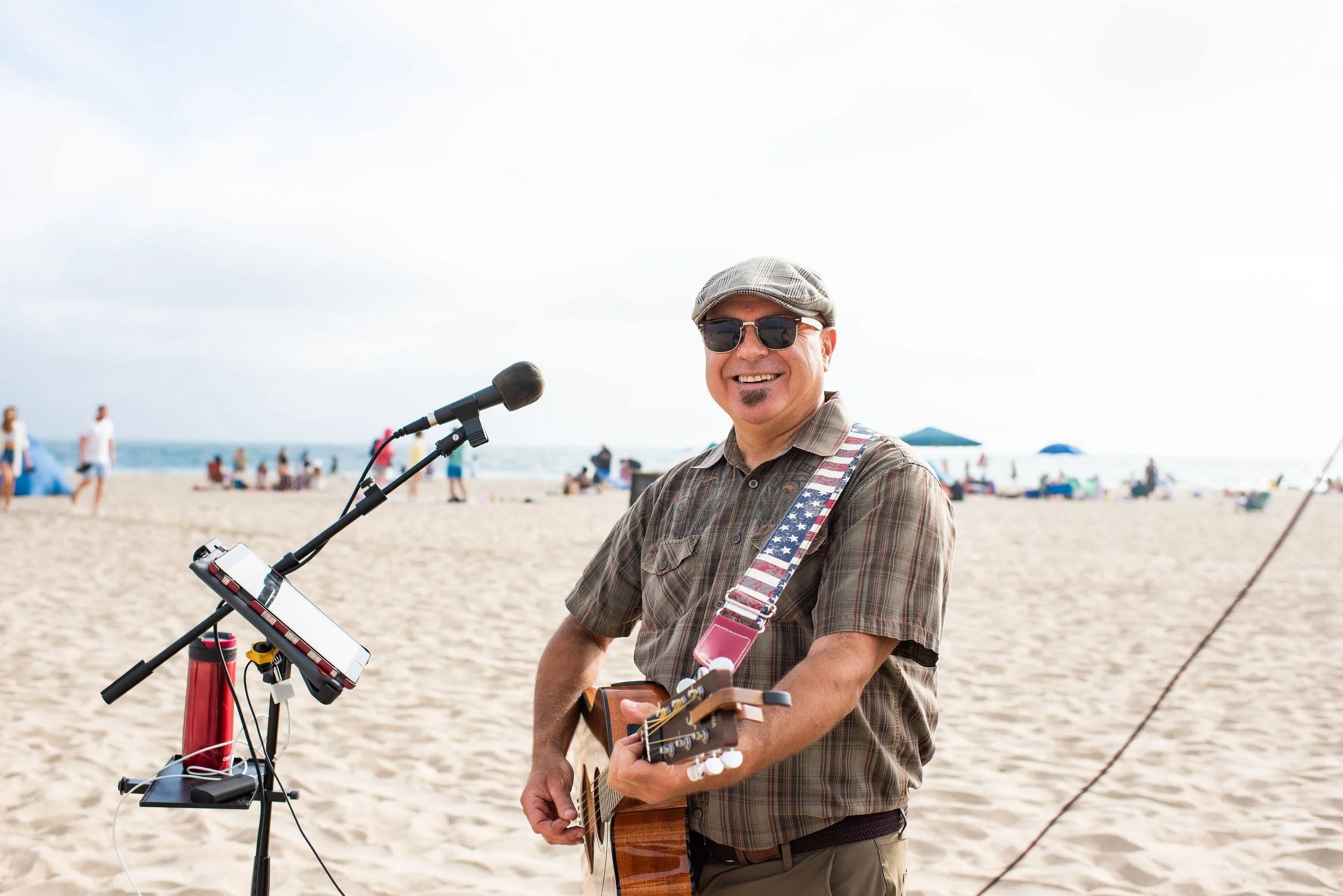 A man wearing sunglasses, a cap, and a plaid shirt smiling and playing an acoustic guitar on a beach with people in the background.