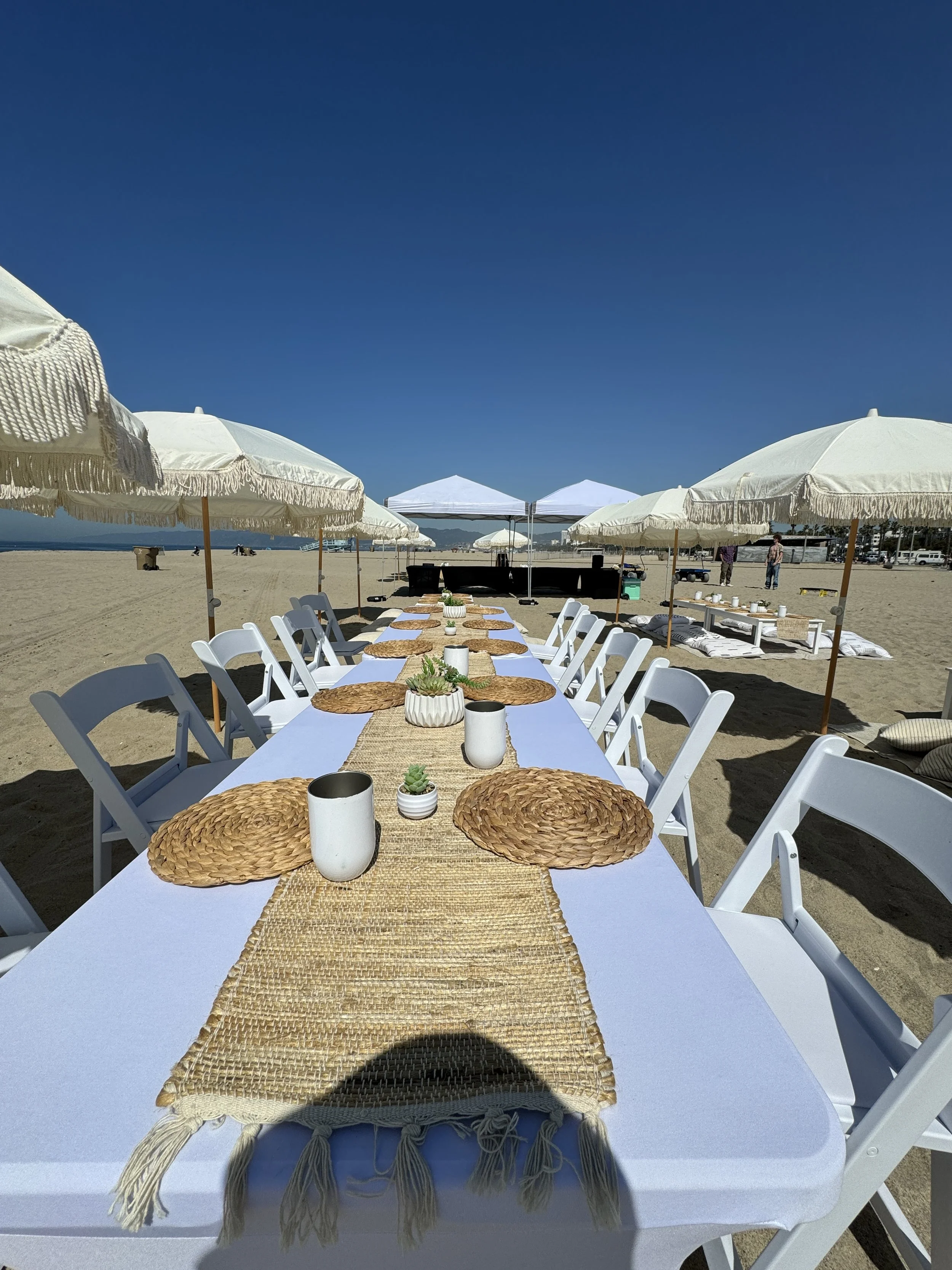 Beach setup with a long white table, woven placemats, white chairs, small potted succulents, and white mugs, shaded by large white umbrellas on sandy beach under a clear blue sky.