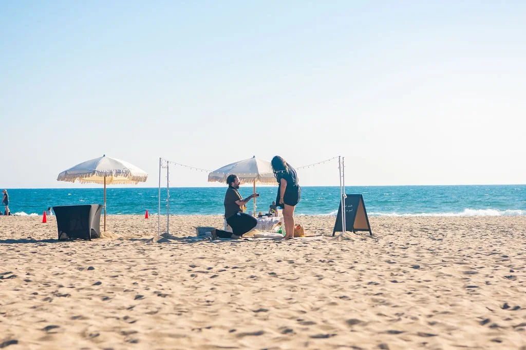Two people at a beach stand under umbrellas, with one kneeling and the other standing, near a small table with a chalkboard sign, on a sandy beach with the ocean in the background.