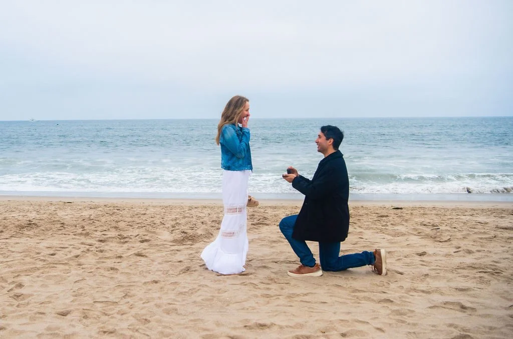 A man is kneeling on one knee on a sandy beach, proposing to a woman, who looks surprised and happy, with the ocean in the background.