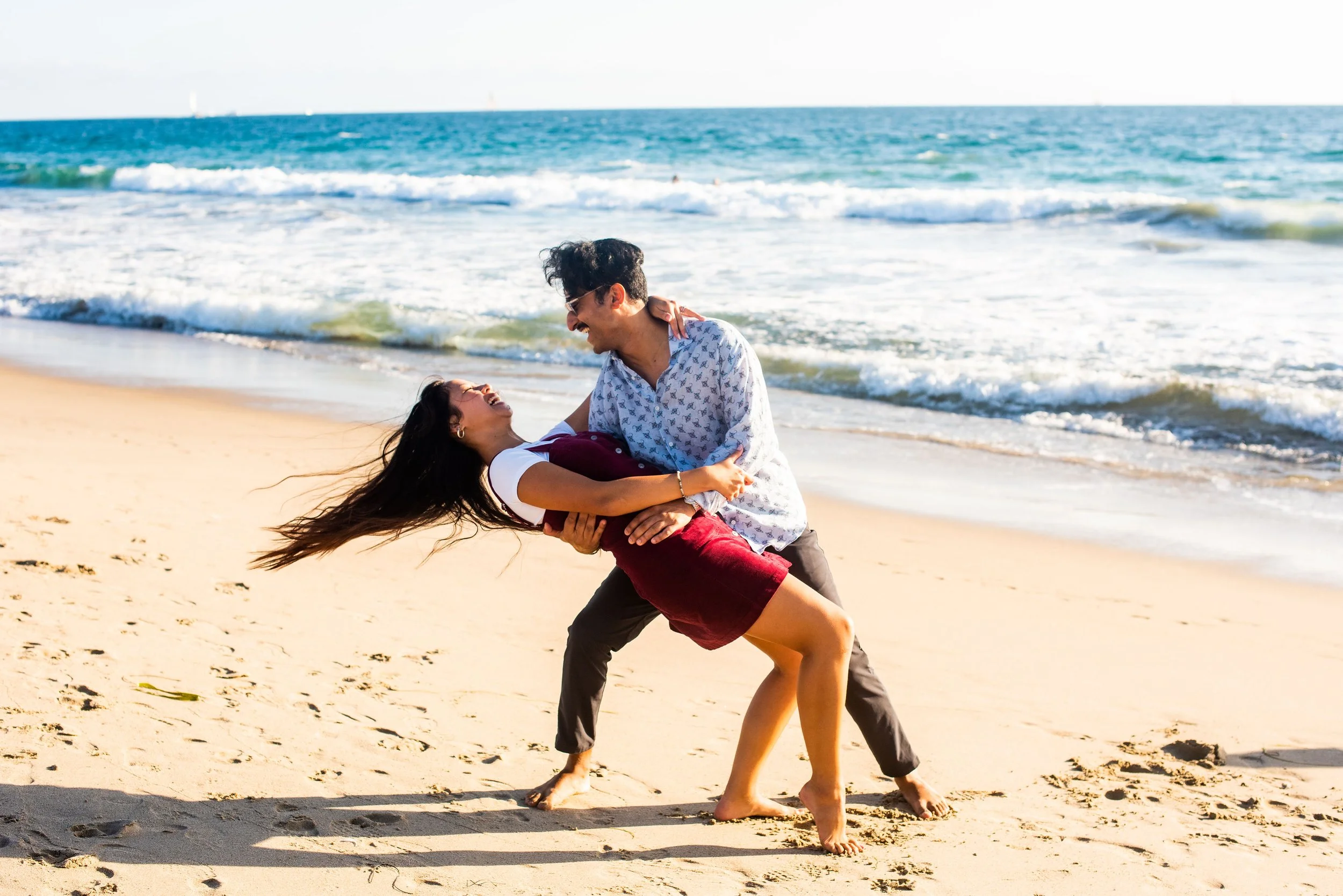 A man and woman laughing and playing on the beach, with the man holding the woman in a dip near the shoreline, ocean waves in the background.