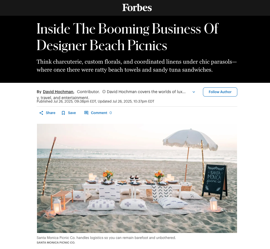 A decorated beach picnic setup with a low table, cushions, flowers, and lanterns on the sand under a white umbrella at Santa Monica Beach.