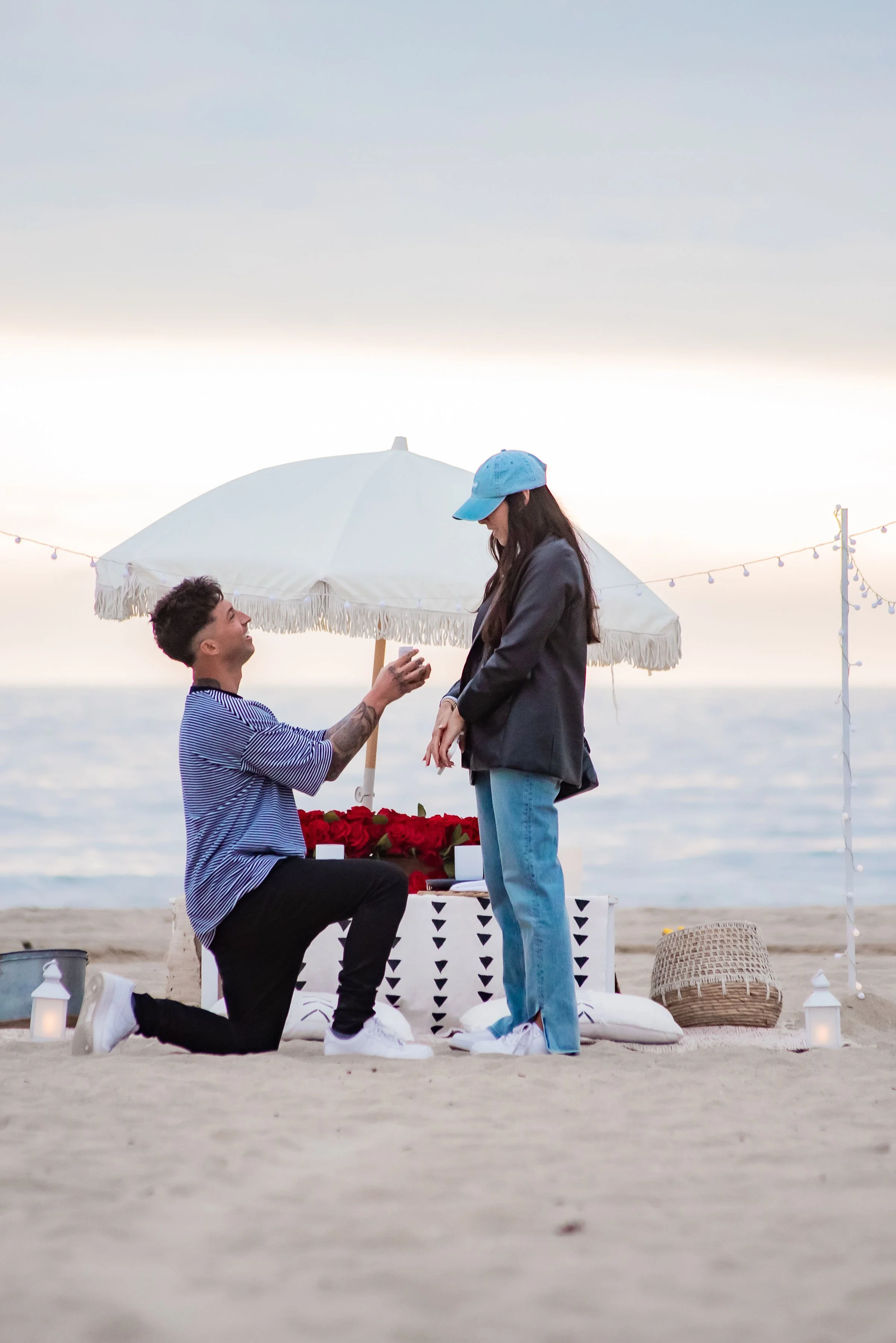 A man proposing marriage to a woman on a beach at sunset, with a decorated setup including an umbrella, roses, candles, and string lights.