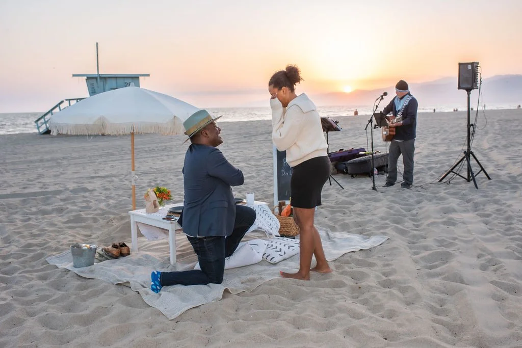 A couple on a beach during sunset, with the man proposing marriage to the woman, while a musician plays guitar nearby.