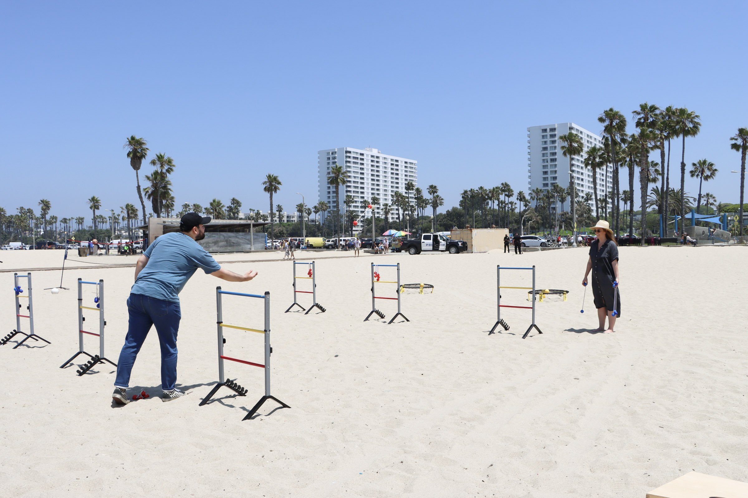 People practicing jump rope exercises on the beach with hurdles set up, tall palm trees, and modern buildings in the background under a clear blue sky.
