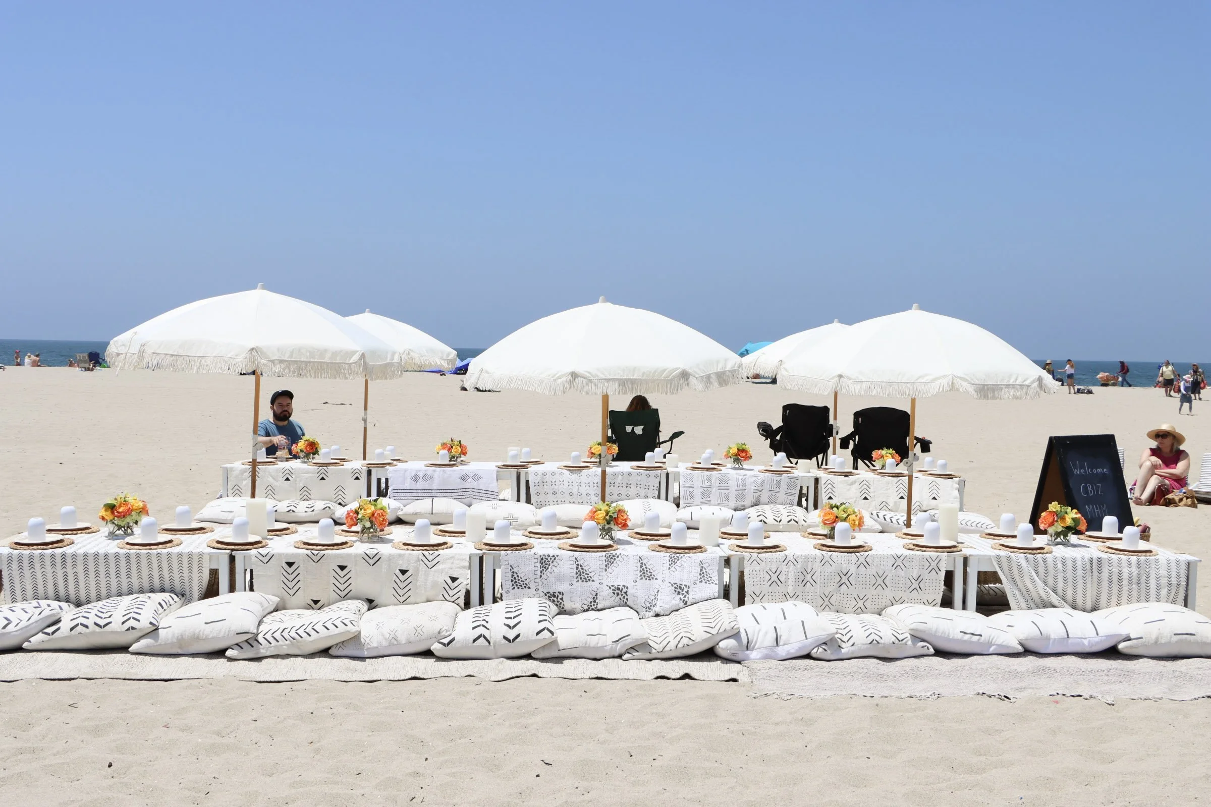 Beach setup with white tables, cushions, and umbrellas for a outdoor event, with people relaxing on the sand and the ocean in the background.
