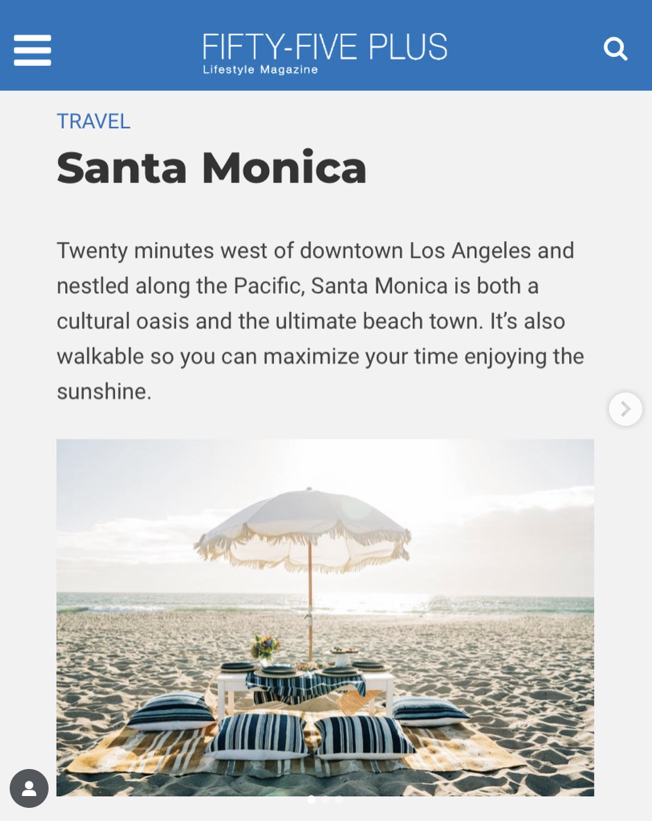 Beach setup with a table, pillows, and a large umbrella on the sandy shore with the ocean in the background, under a partly cloudy sky.