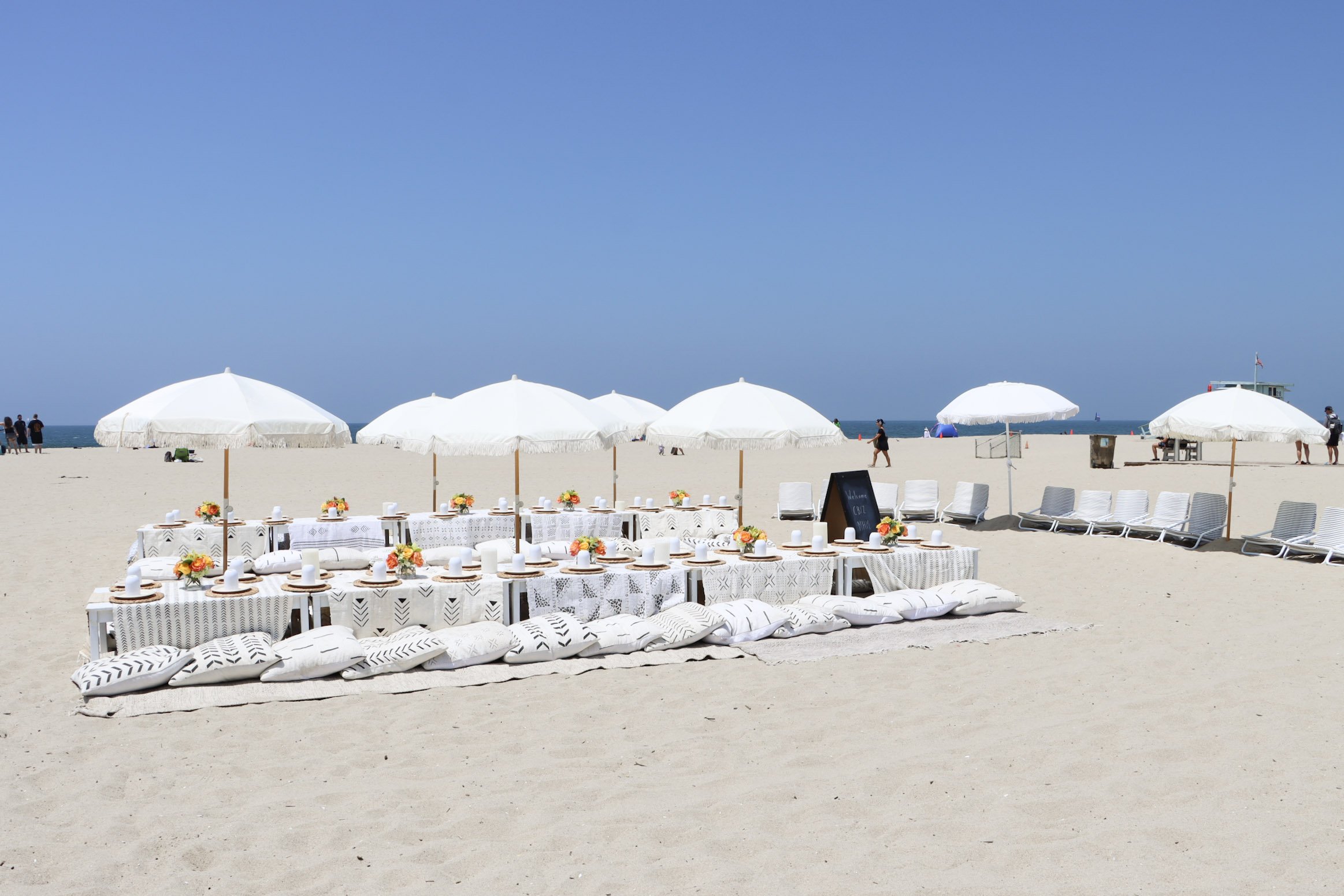 Beach setup with low tables, umbrellas, and cushions for a gathering, overlooking the ocean on a sunny day.