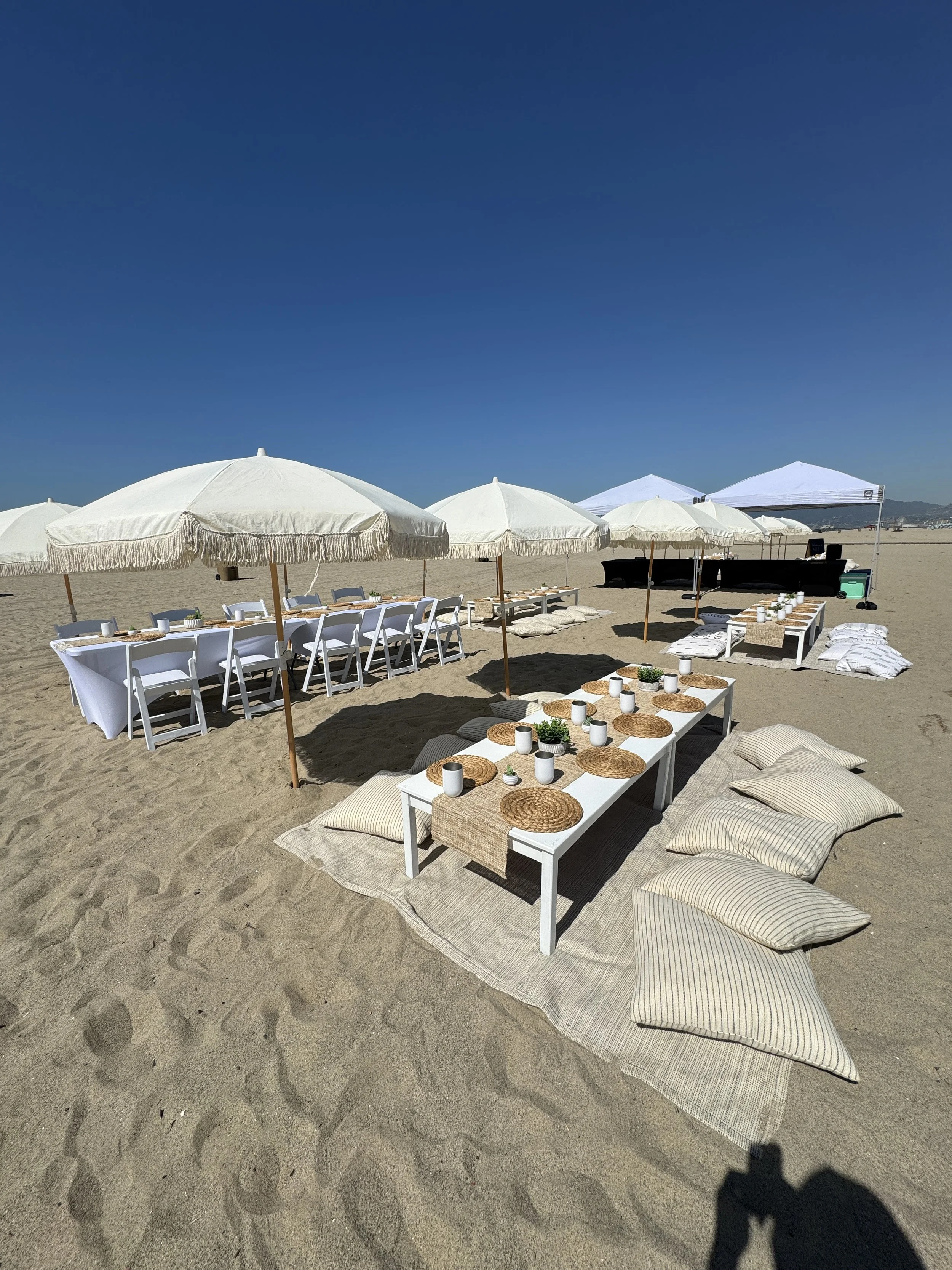 Beach setup with white umbrellas, tables, cushions, and tableware for outdoor dining or event, under a clear blue sky.