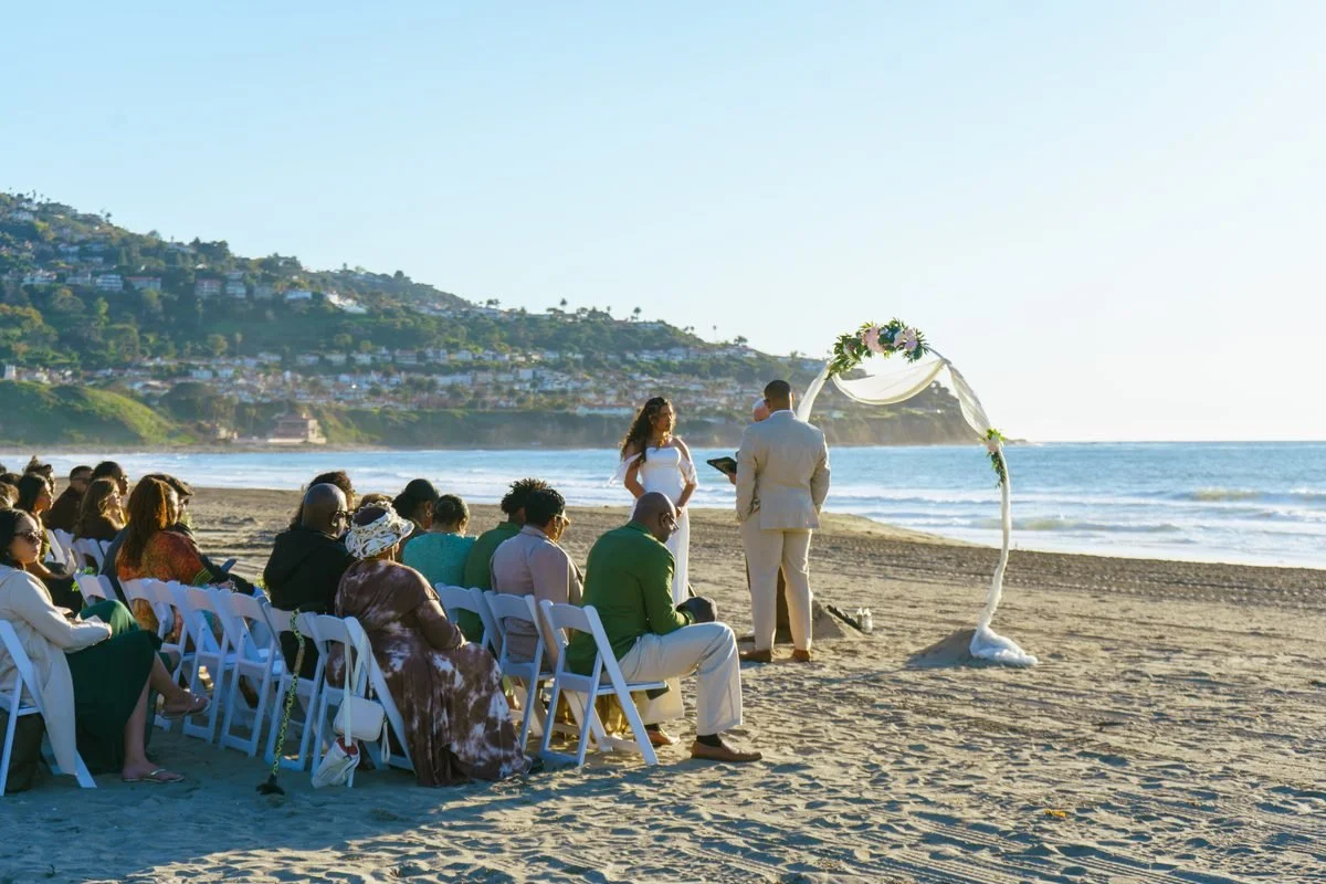 Sets of white chairs arranged in rows on a sandy beach, facing a decorated wedding arch with orange and red flowers, with the ocean and sky in the background.
