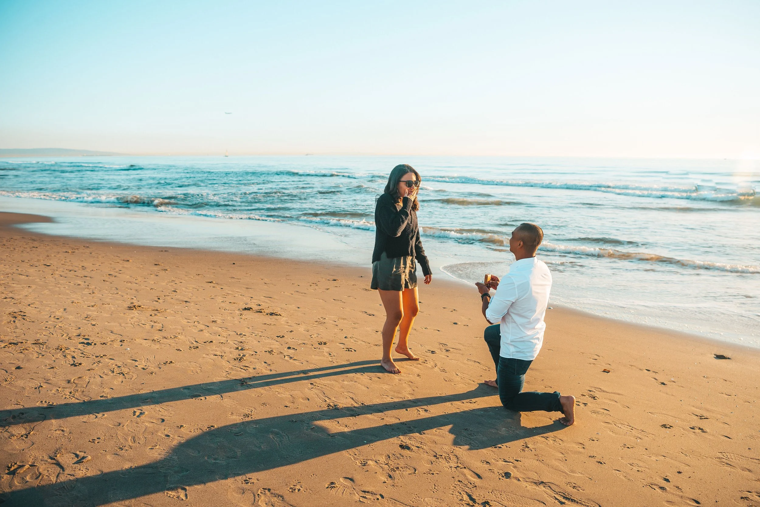 A man on one knee proposing to a woman on a sandy beach at sunset, with the ocean in the background.