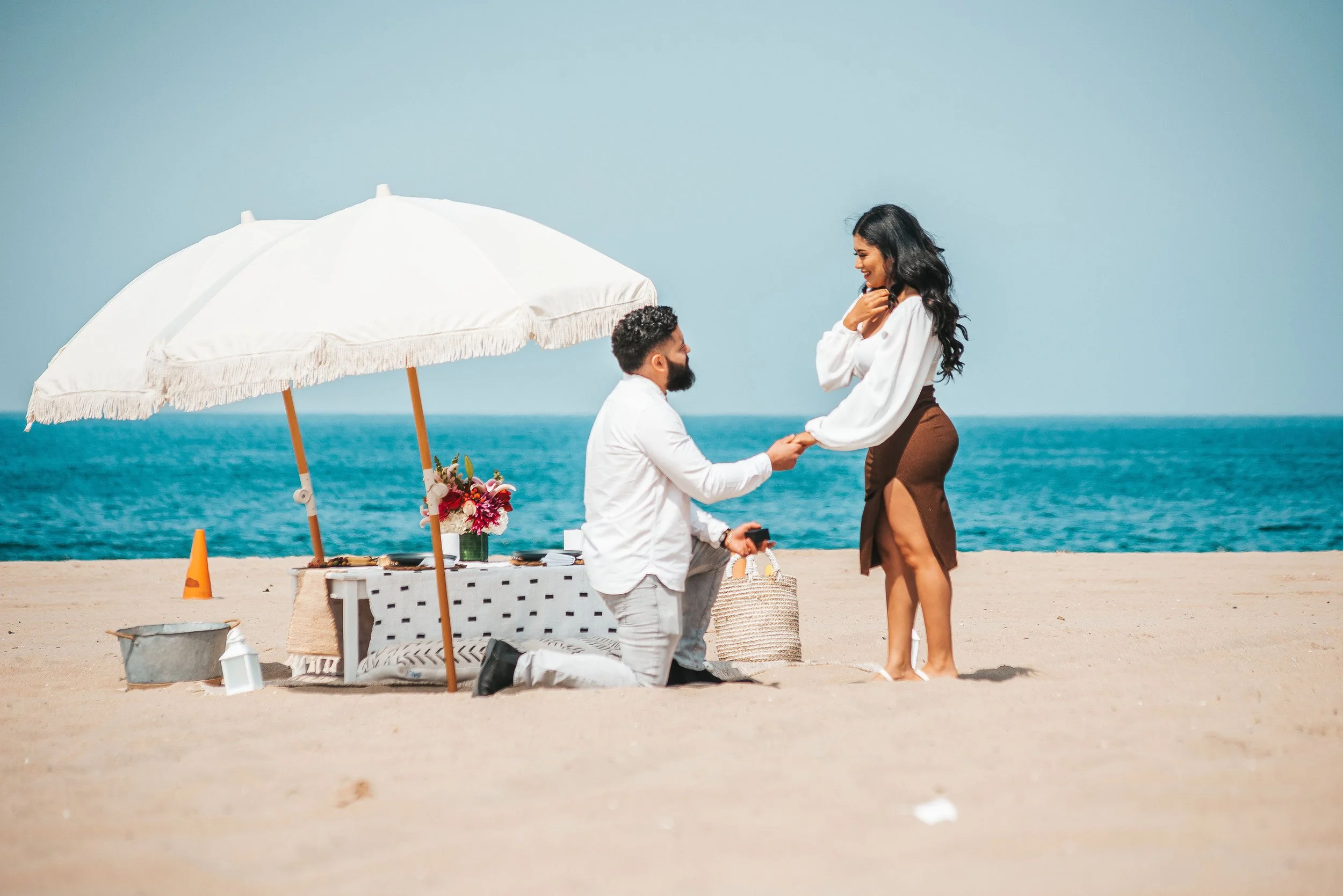 A man proposing marriage to a woman on a beach, with ocean in the background, under a white umbrella, with a small table and flowers nearby.